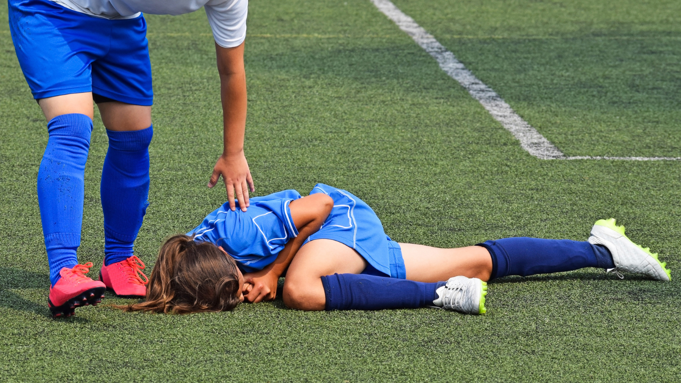 Soccer player in blue uniform injured on field, teammate offering assistance.