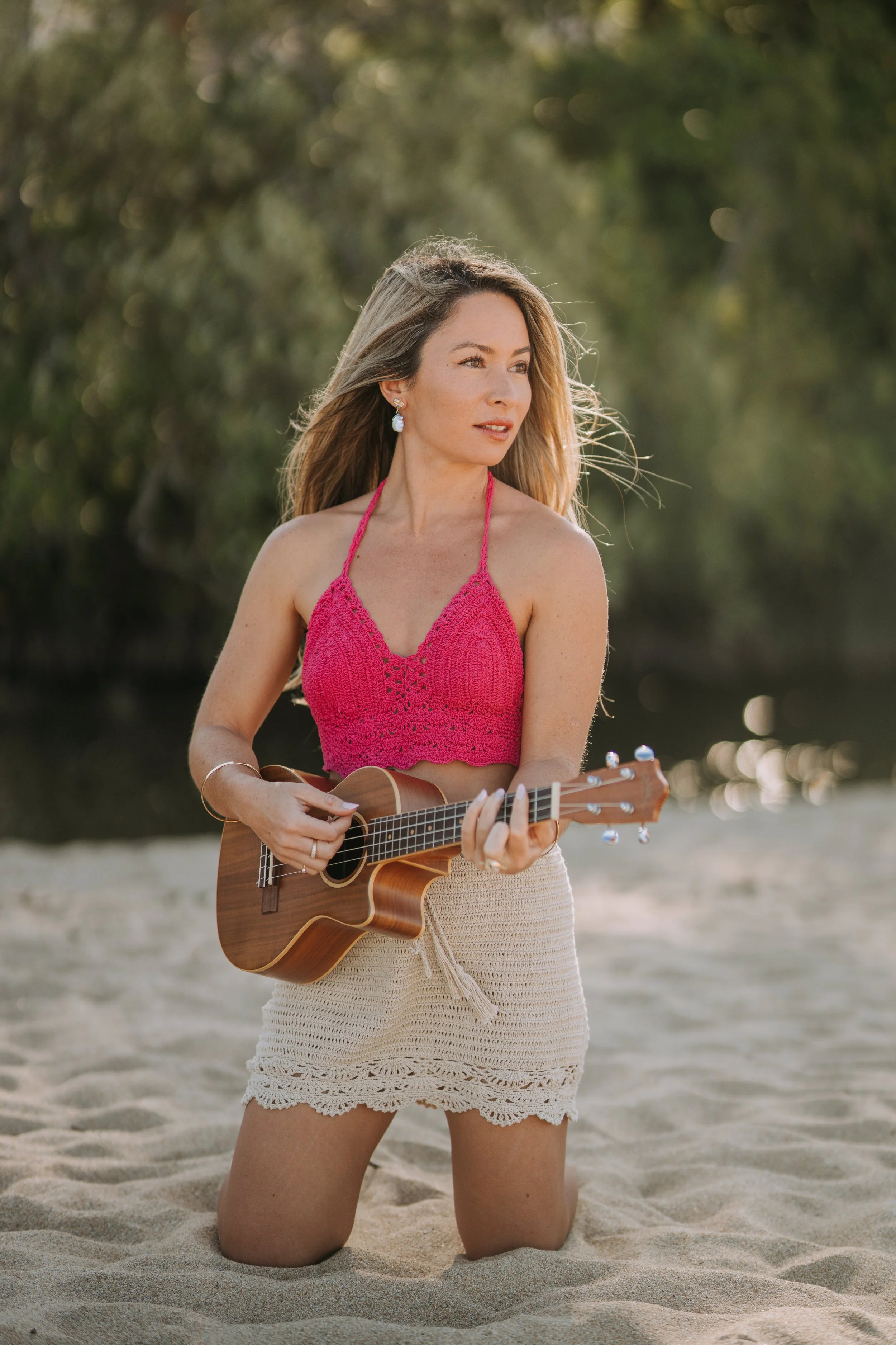 A woman kneeling on sandy ground outdoors, playing a ukulele, with trees in the blurred background. She is wearing a pink crochet top and a beige skirt.