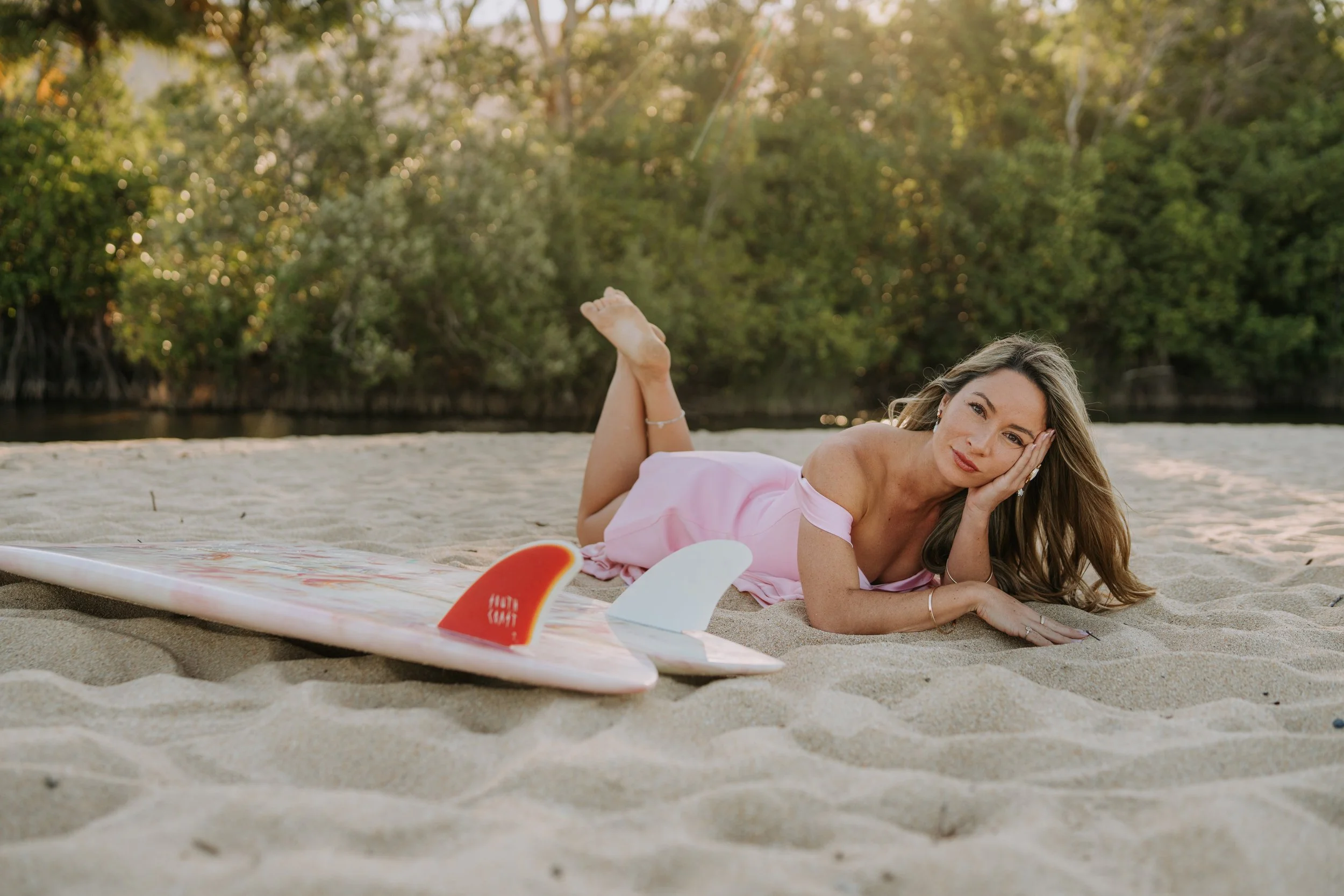 A woman lying on a sandy beach during sunset, wearing a pink dress, with a surfboard nearby, and trees in the background.