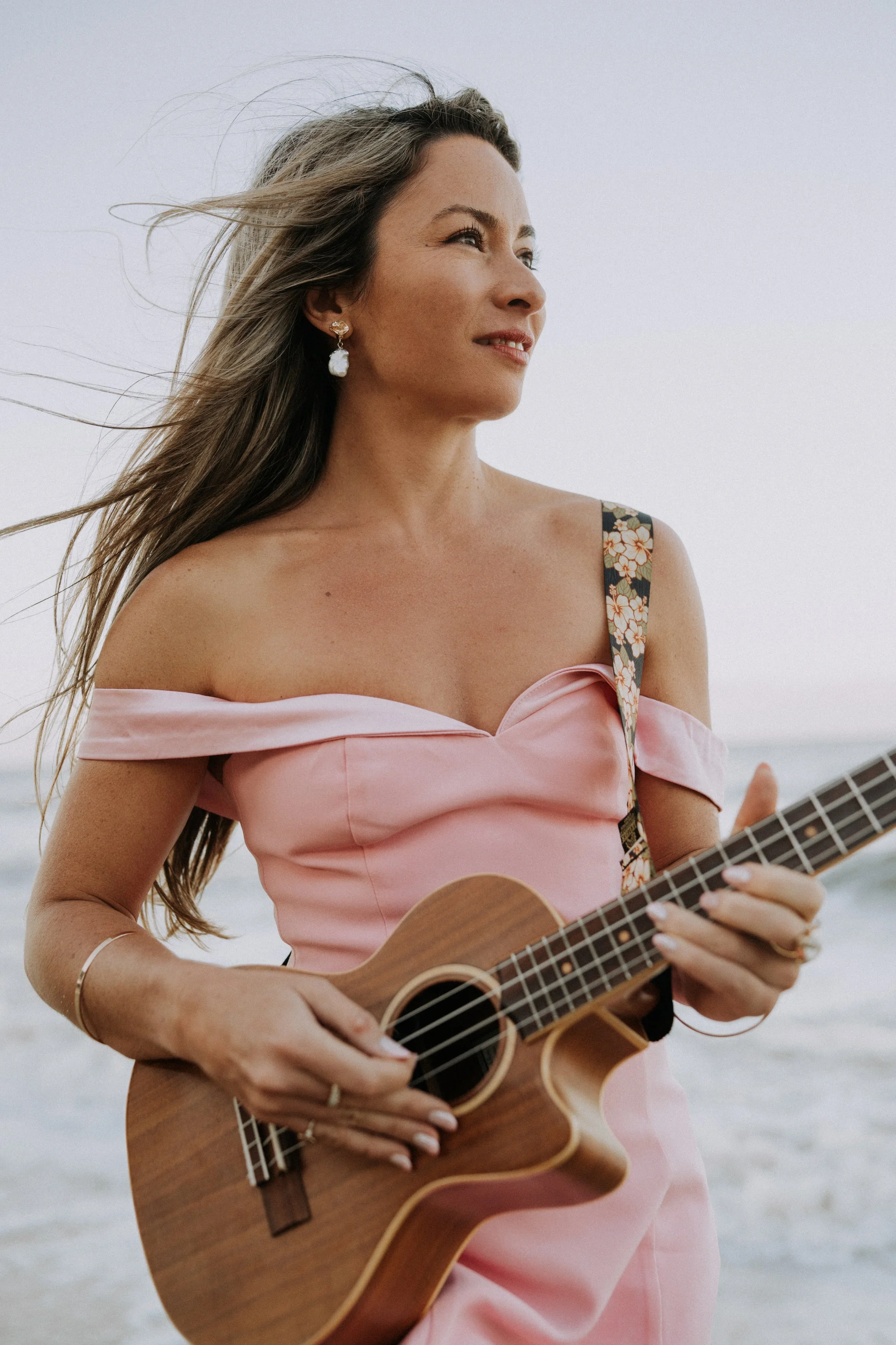 A woman with long, wind-blown hair standing on a beach, playing a ukulele, wearing a pink off-shoulder dress with floral strap, earrings, and a bracelet.