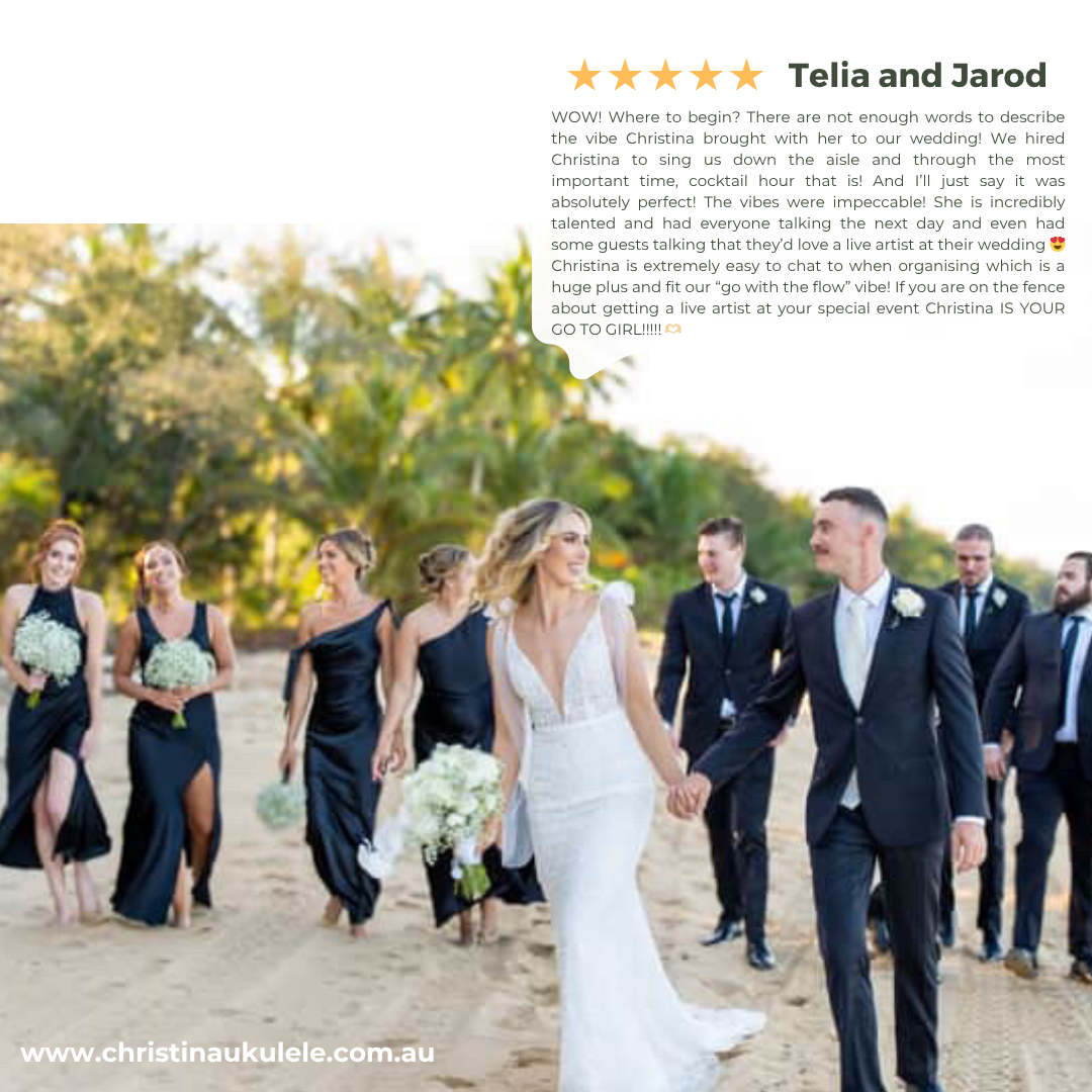 A wedding party walking on a sandy beach with a bride in a white gown holding a bouquet and groom in a dark suit holding hands, surrounded by bridesmaids in navy dresses and groomsmen in dark suits, with trees in the background.