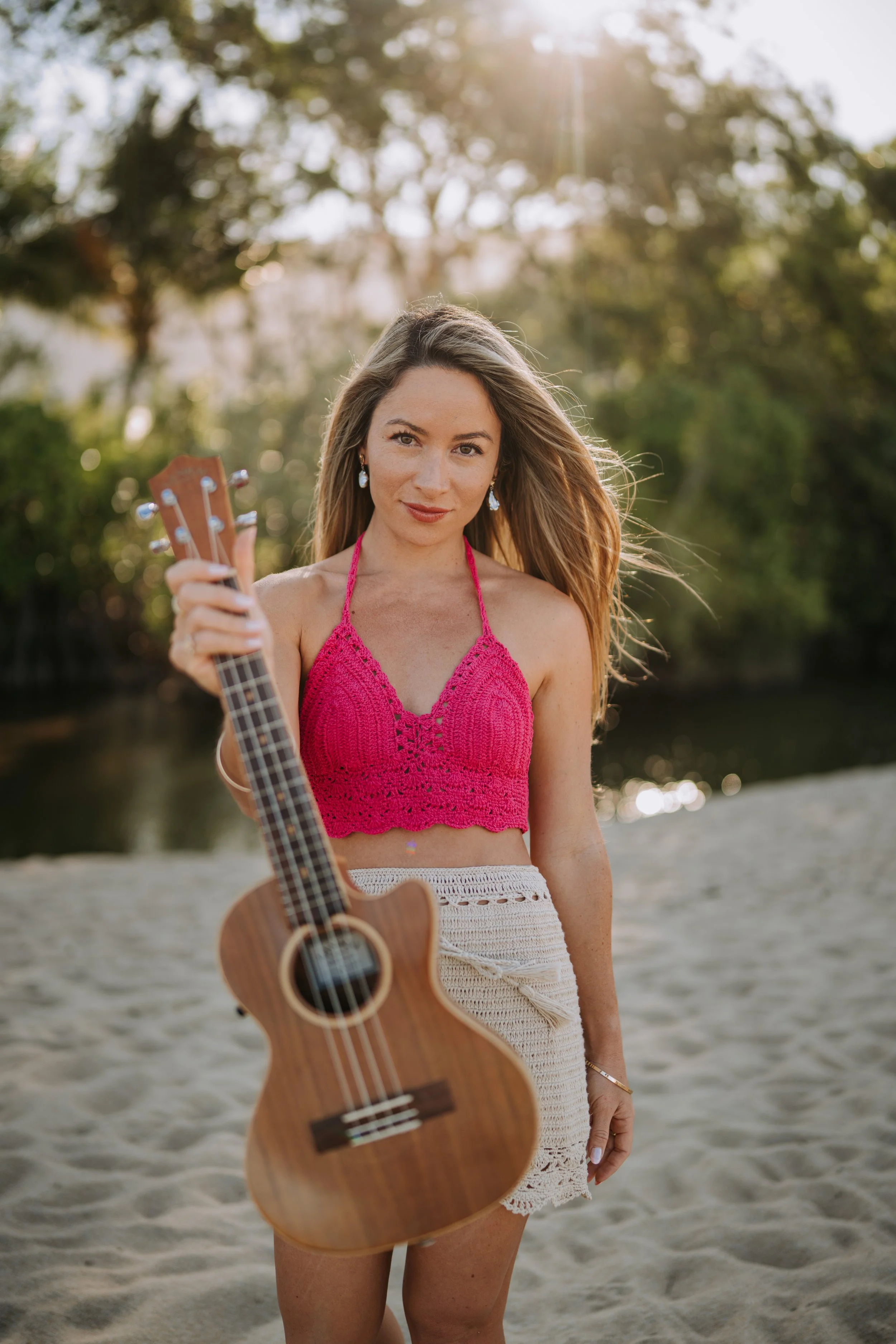 A woman holding a ukulele standing on a sandy beach with trees and water in the background during sunset.