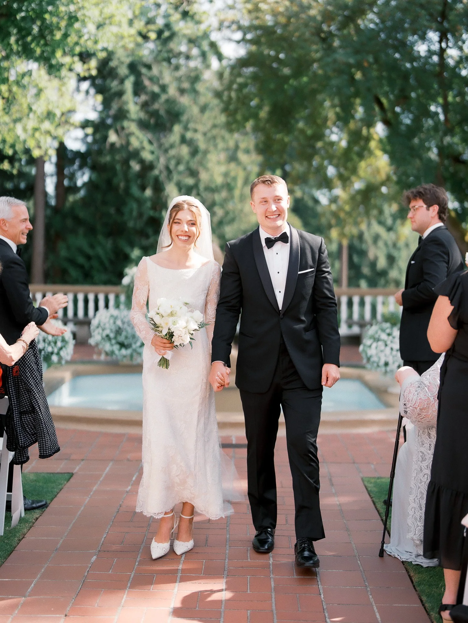 Bride and groom walking hand in hand at their outdoor wedding ceremony, smiling, with guests clapping and celebrating around them.