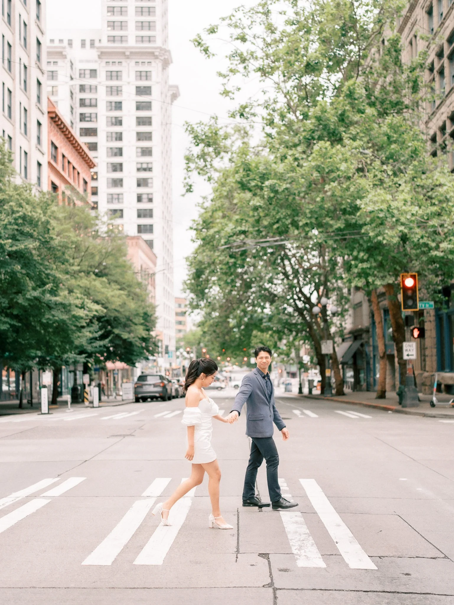 A young woman and man walking across a city street at a crosswalk holding hands, with tall buildings and green trees in the background.
