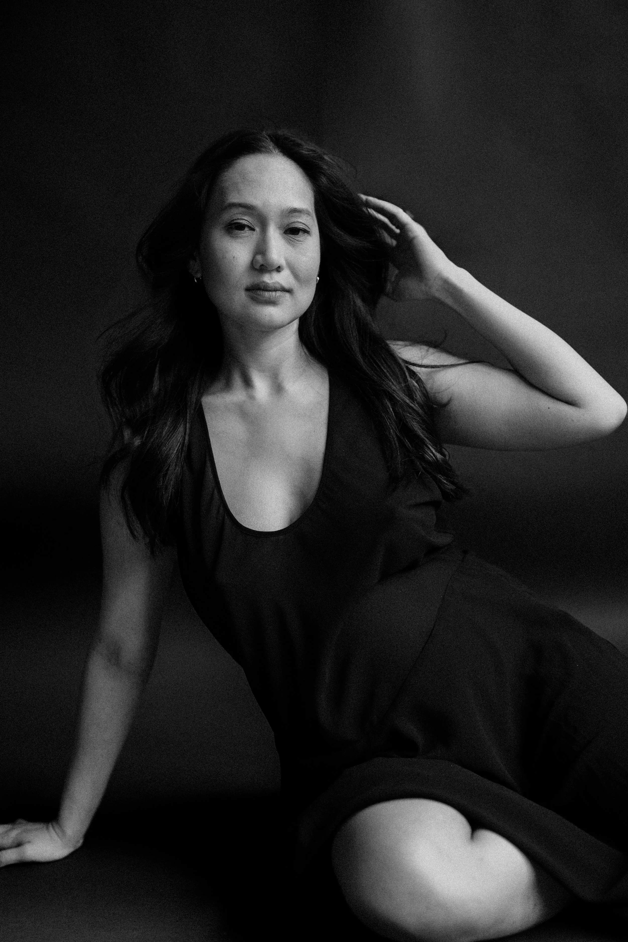 Black and white portrait of a woman with long hair, wearing a sleeveless top, sitting on the floor against a dark background, gazing at the camera with one hand touching her hair.