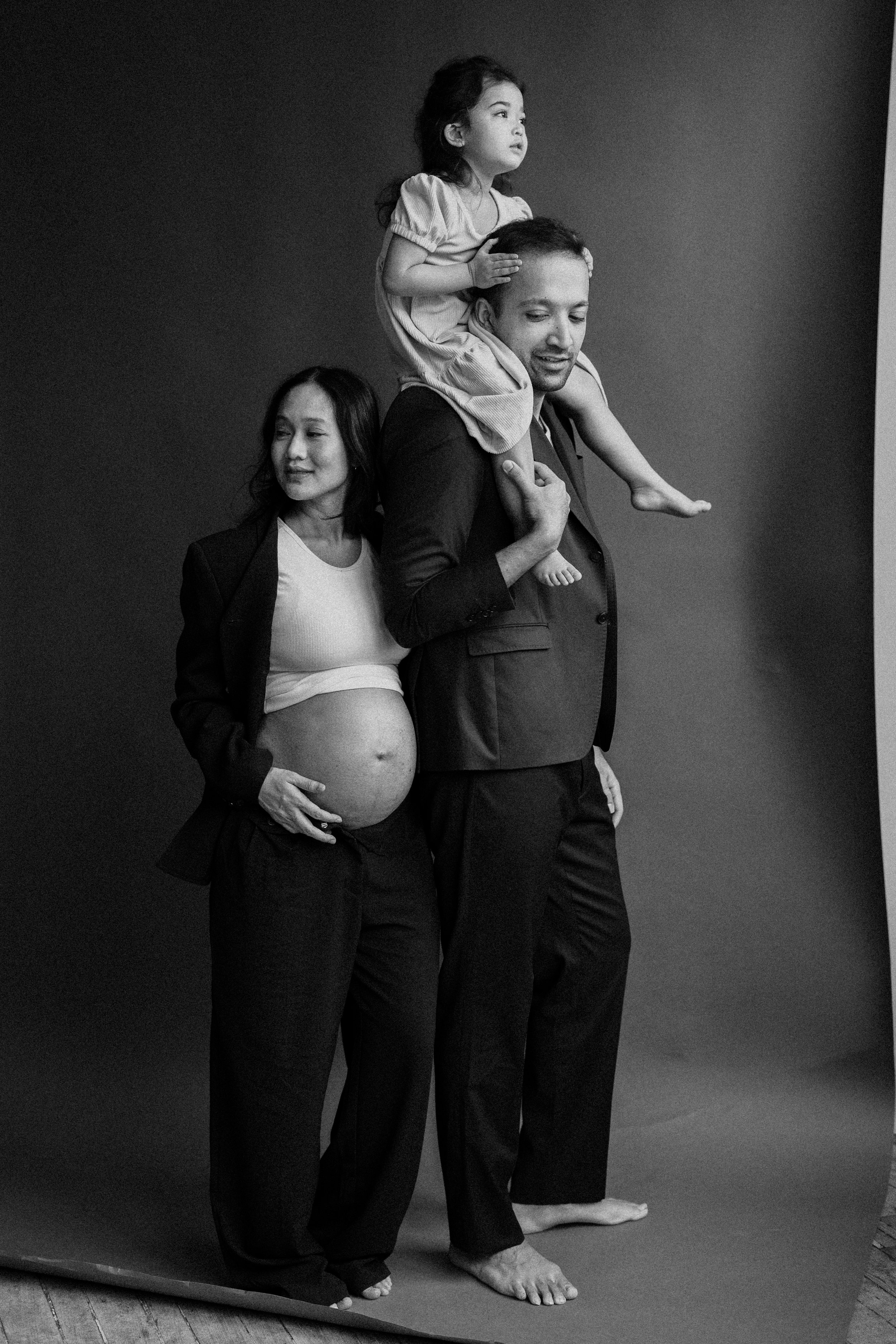 A black-and-white photo of three women, one pregnant and two children, standing against a plain background. The woman on the left is pregnant and touching her belly. The woman in the middle is carrying a young girl on her shoulders, who is looking to