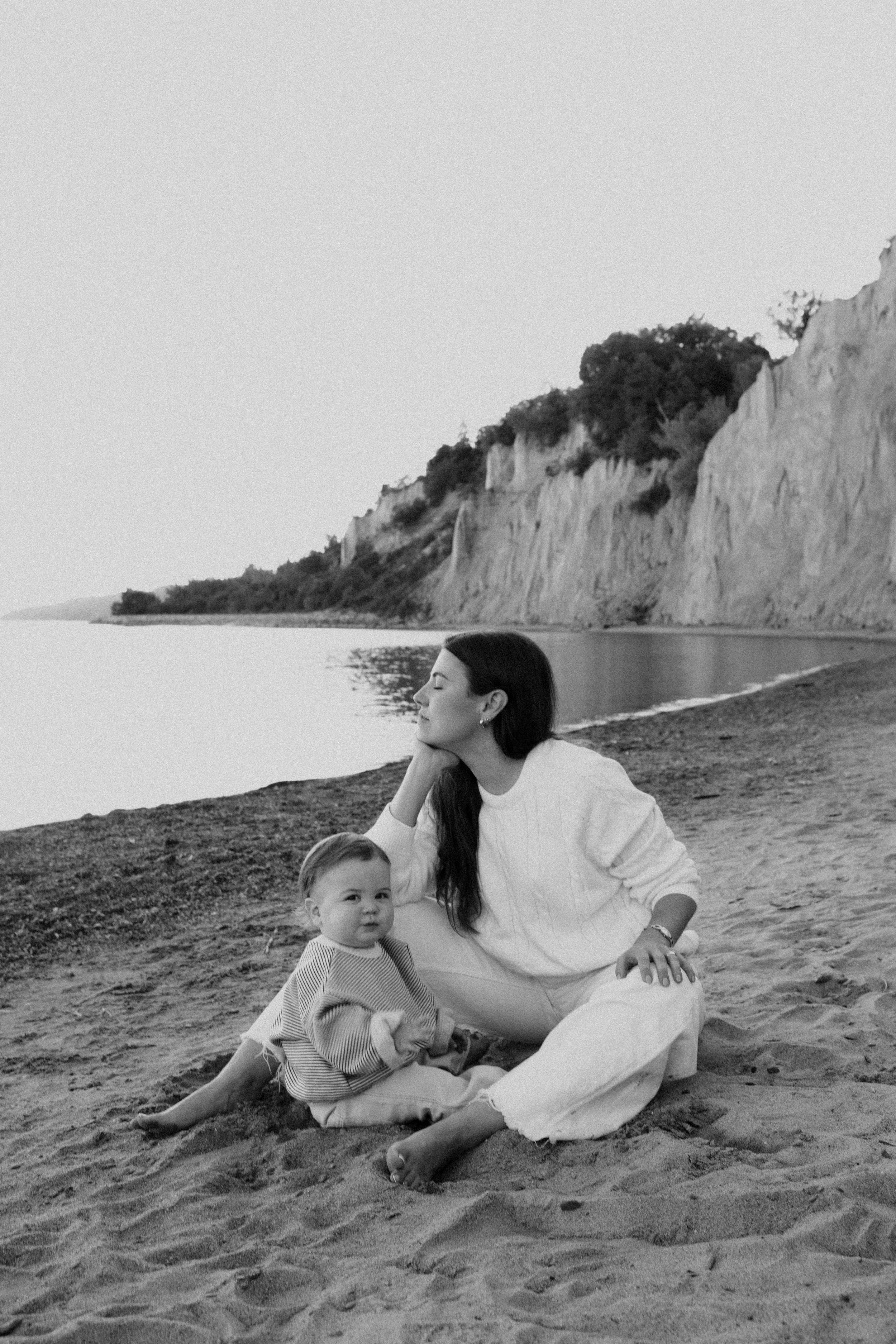 A woman and a young girl sitting on a sandy beach near water with cliffs in the background, black and white photograph.