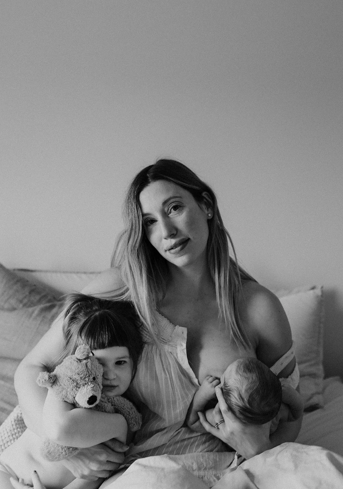 A black and white photo of a woman breastfeeding a baby, holding a young girl with a stuffed animal, sitting on a bed with pillows in the background.