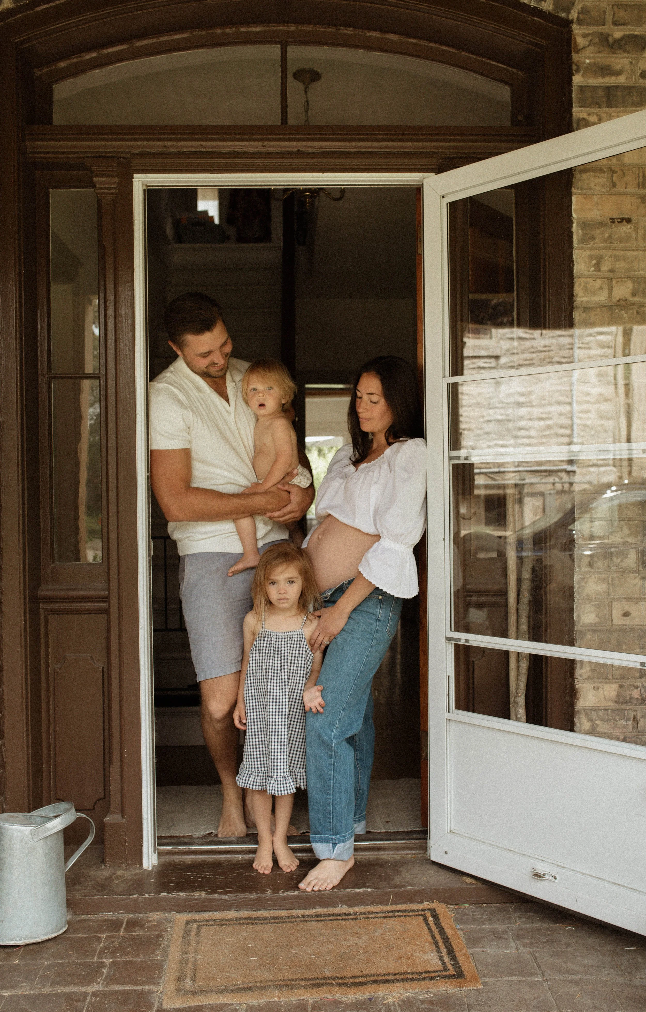 Family standing on front porch, including a pregnant woman, man holding a toddler, and a young girl in a checkered dress.