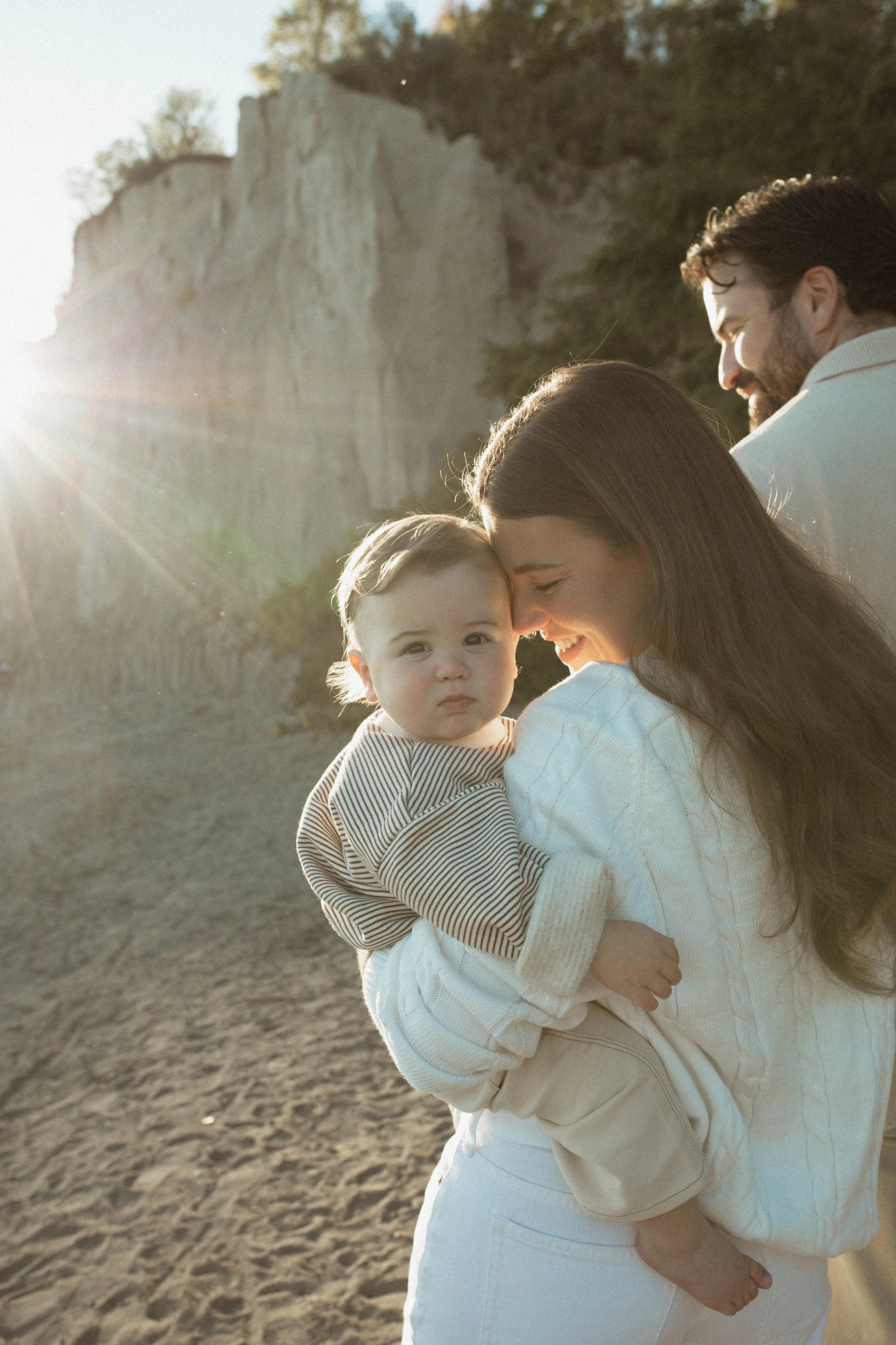 A family with a woman, man, and young child on a beach with cliffs in the background. The woman is holding the child, and the man is smiling nearby. The sun is setting or rising, creating a warm glow.