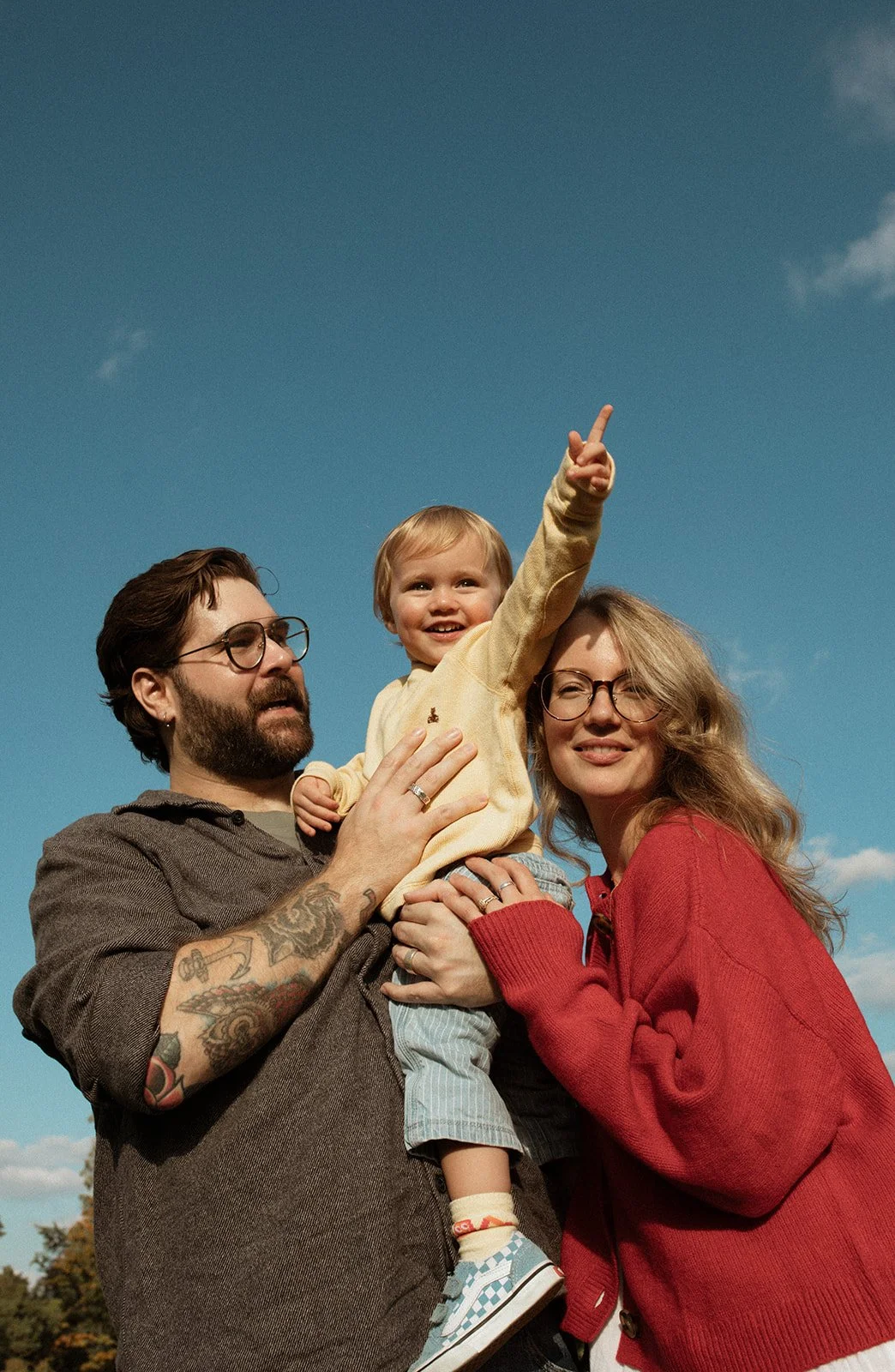 A family of three, including a man, woman, and young boy, standing outdoors under a clear blue sky. The man is holding the boy, who is pointing upward, and both parents are smiling.