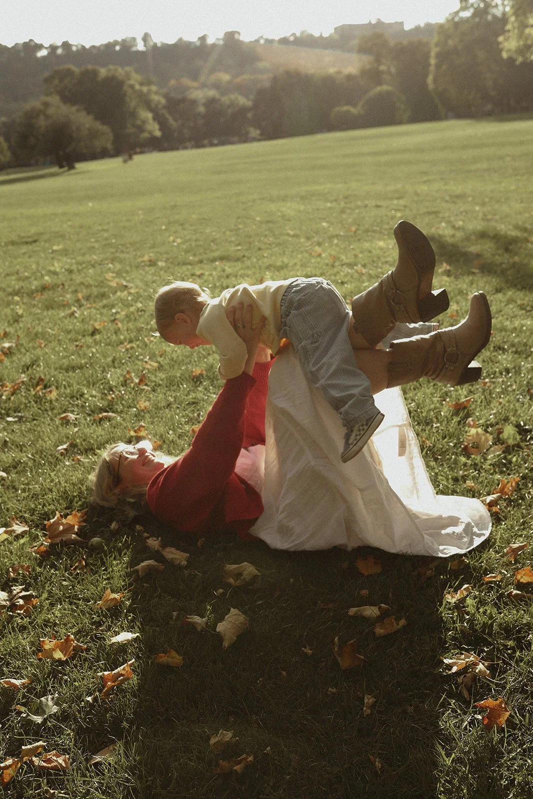 An older woman with glasses and blonde hair lying on the grass, playing with a young boy. The boy is in mid-air, facing the woman, as she lifts him up. They are outdoors in a park with trees and hills in the background.