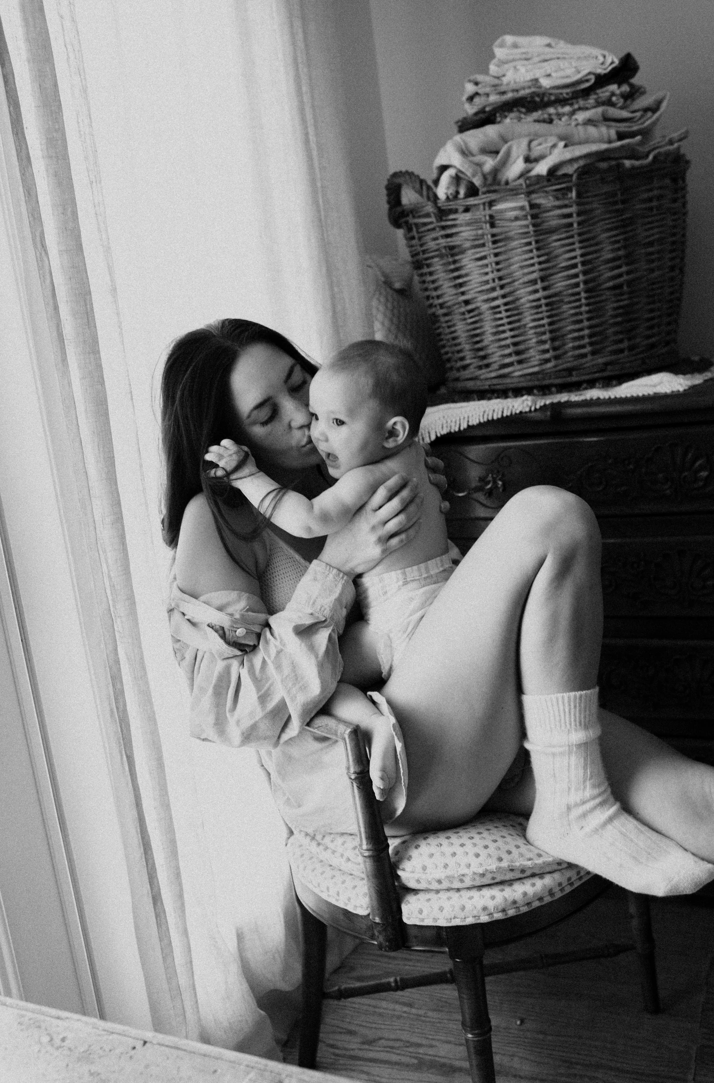 A woman with long dark hair holding a young child on her lap, sitting on a chair, near a window with curtains. The woman appears to be giving the child a kiss on the cheek. In the background, there is a basket of laundry on top of a dresser.
