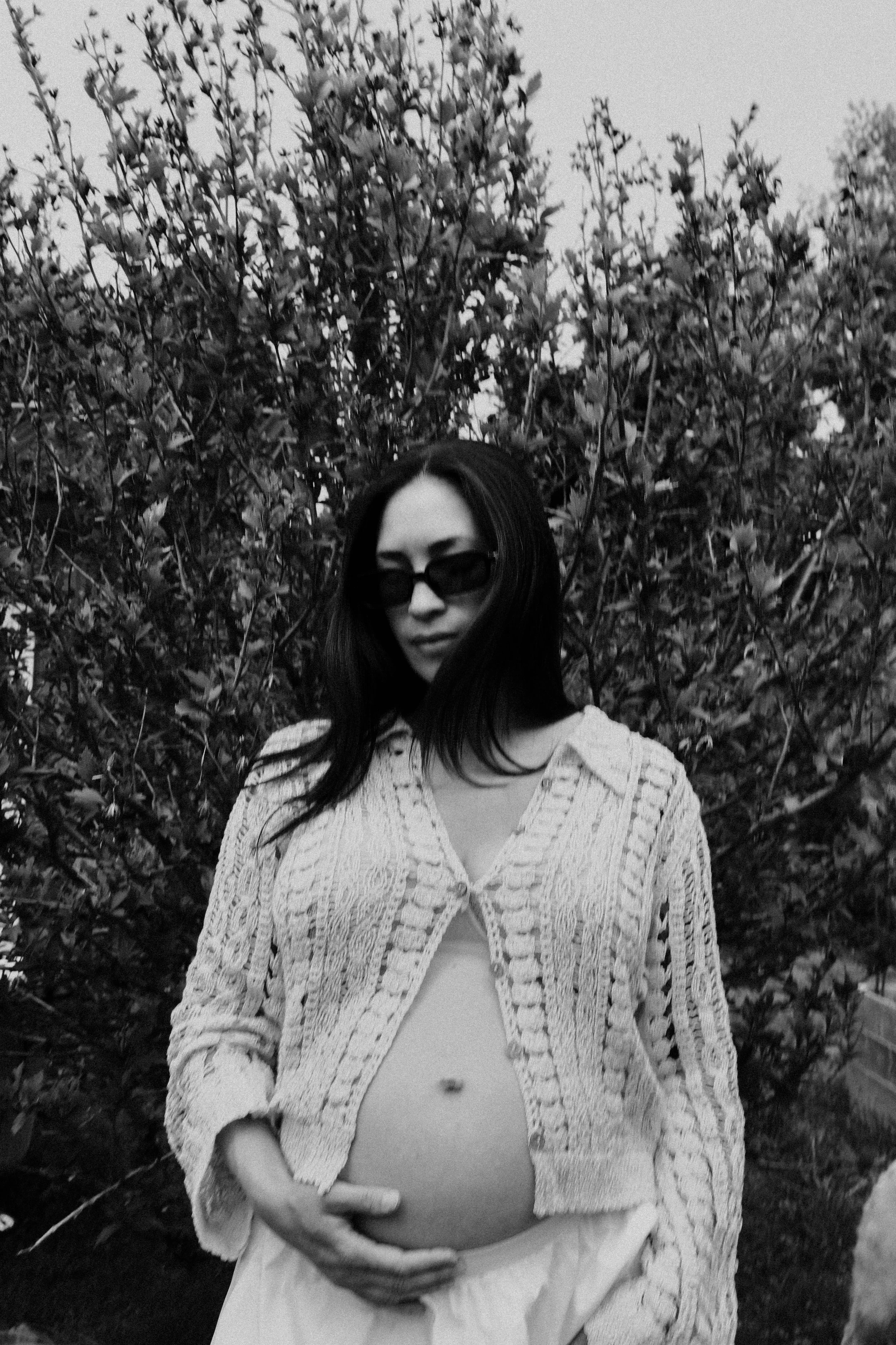 A woman with long dark hair wearing sunglasses and a knitted cardigan, holding her pregnant belly outdoors in front of dense bushes, in black and white.
