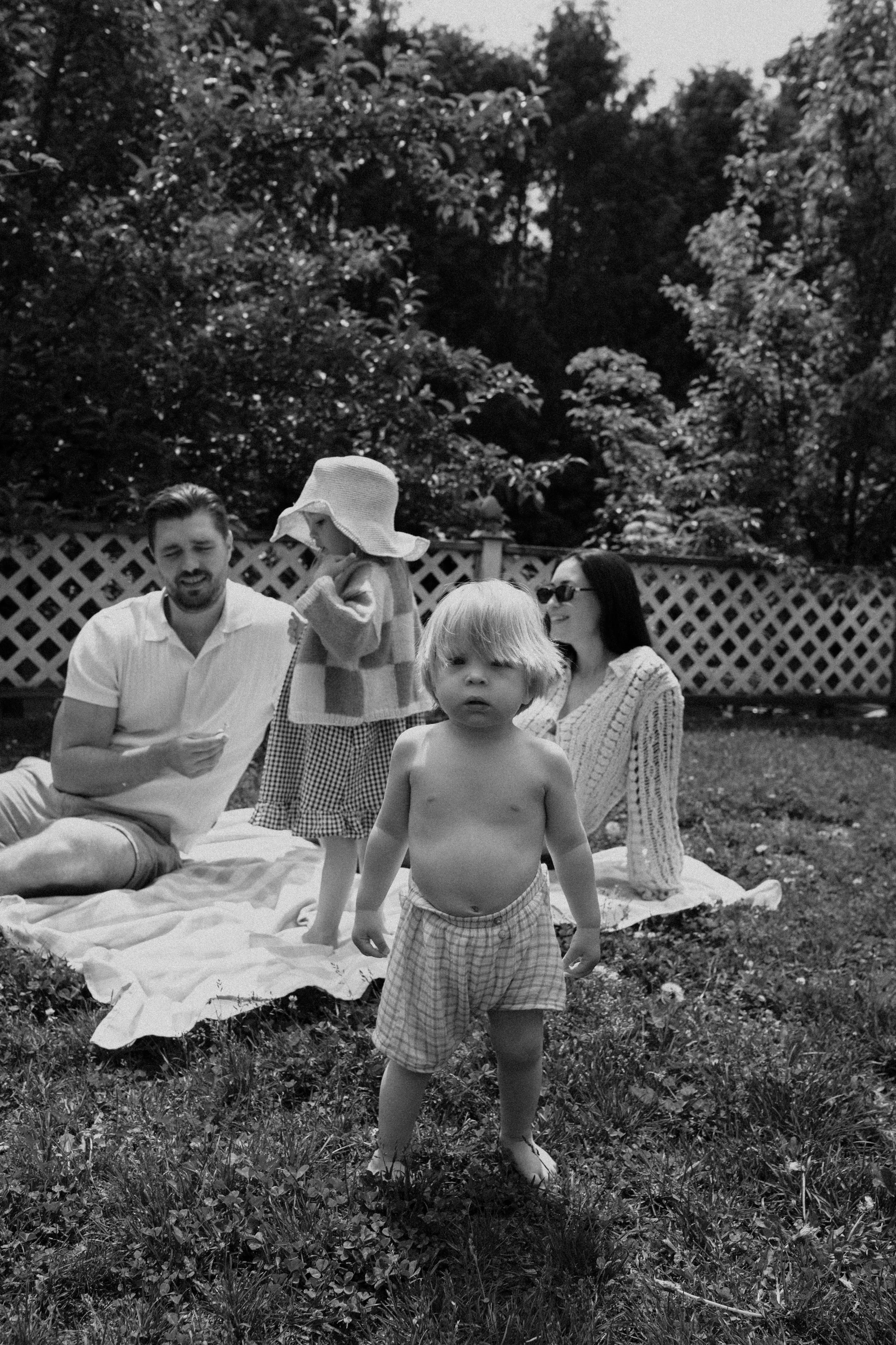 A black and white photo of a family enjoying a picnic outdoors on a blanket in a yard with trees and a wooden fence. Two children, one shirtless boy in plaid shorts standing in the foreground and a girl wearing a hat and checkered dress, are present.