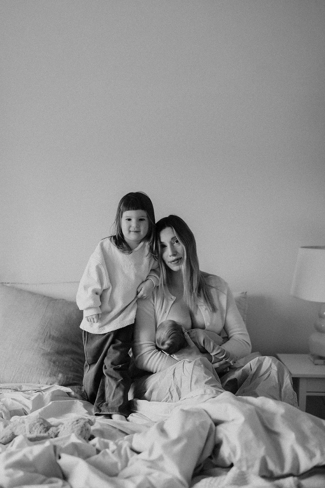 A woman with a new born baby and a young girl sitting on a bed, all looking at the camera in a monochrome photo.