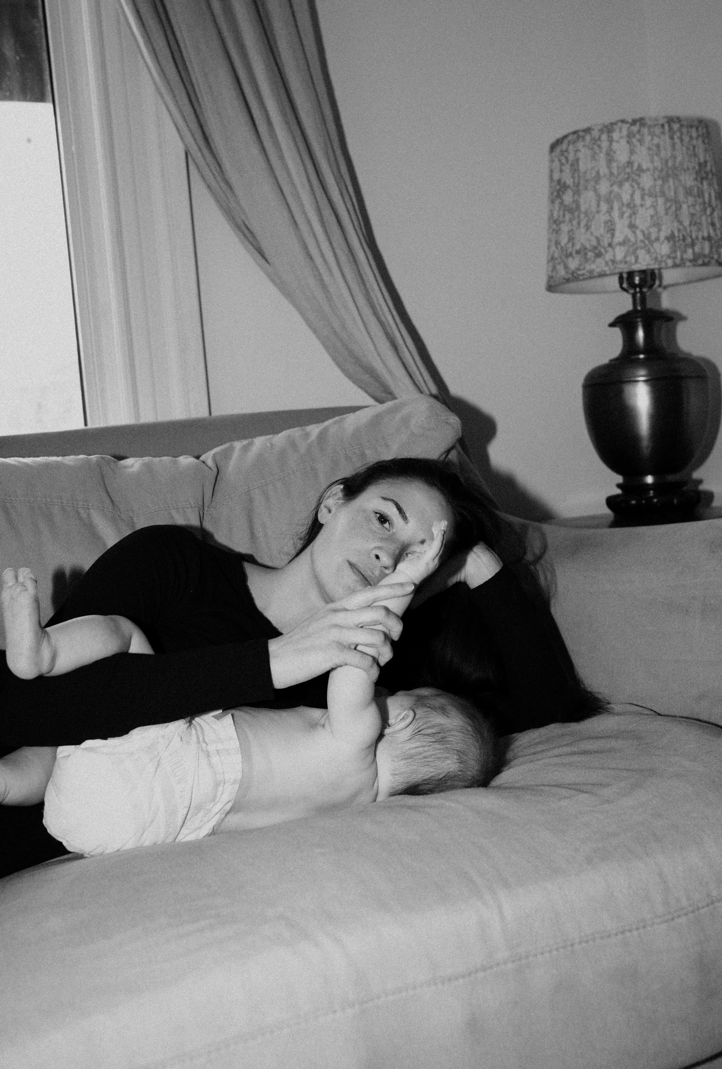 A woman lying on a bed with a baby, holding a finger to her lips for a secret gesture, in a cozy bedroom setting, black and white photograph.