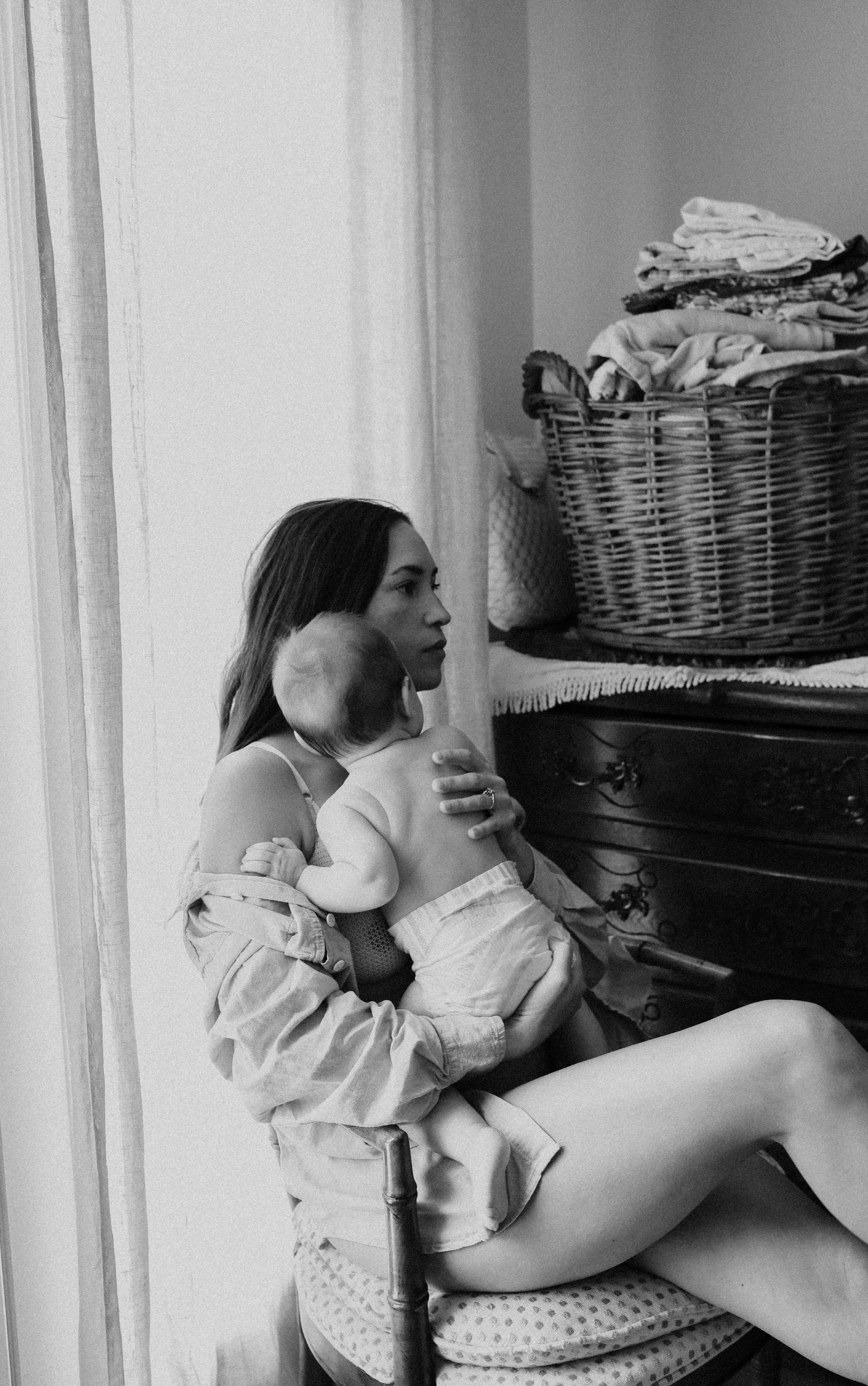A woman sitting on a chair holding a baby close to her chest, with a laundry basket filled with clothes nearby.