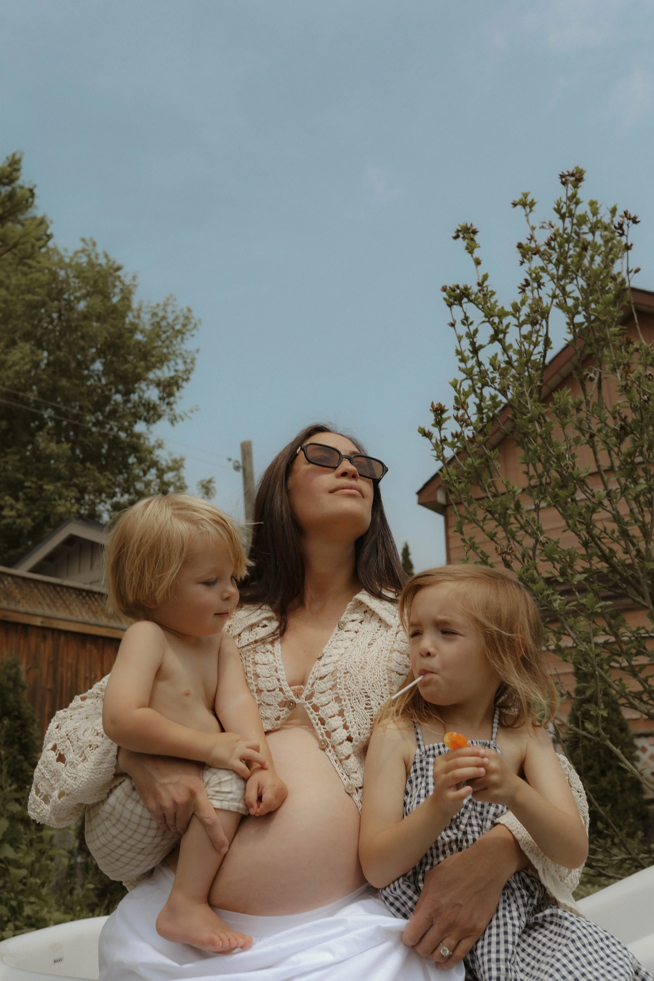 A pregnant woman wearing sunglasses sitting outside with two small children, one child sitting on her lap and the other holding a lollipop, in a backyard.