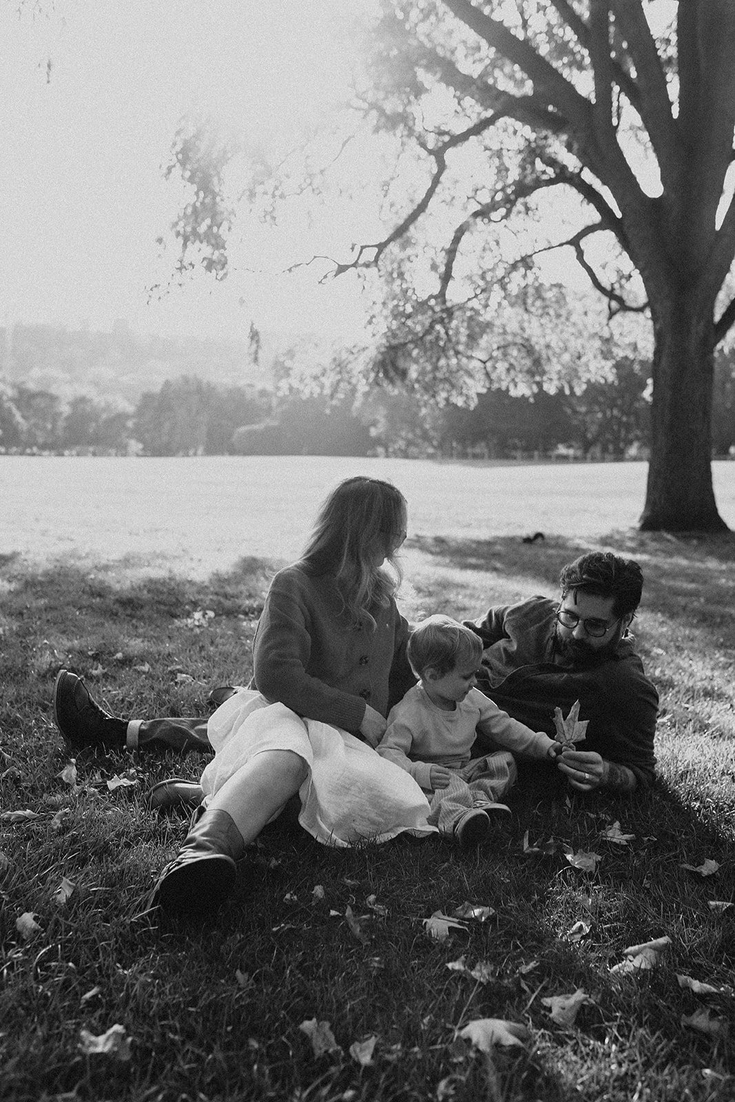 A black and white photo of a family of three sitting on grass under a large tree in a park. The man and woman are sitting facing each other with a small child between them. The man is holding a leaf and the woman appears to be looking at the child, w