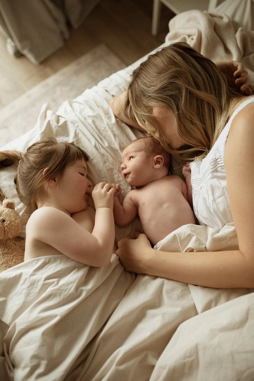 A woman, a young girl, and a baby lying on a bed, holding hands and gazing at each other, all with light skin and surrounded by white bedding.
