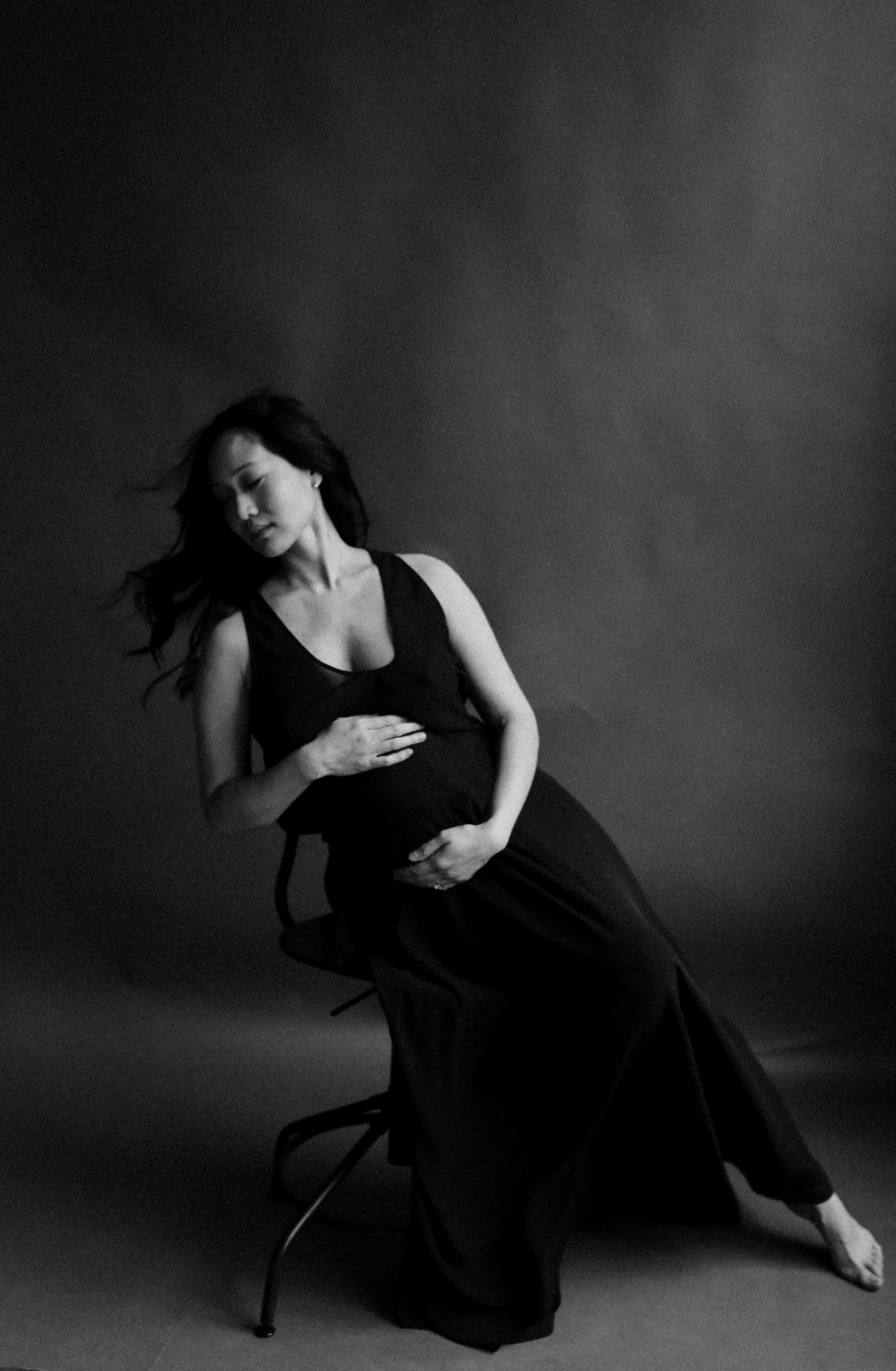 A pregnant woman in a black dress sitting on a chair against a dark background, with her eyes closed and hands resting on her belly.