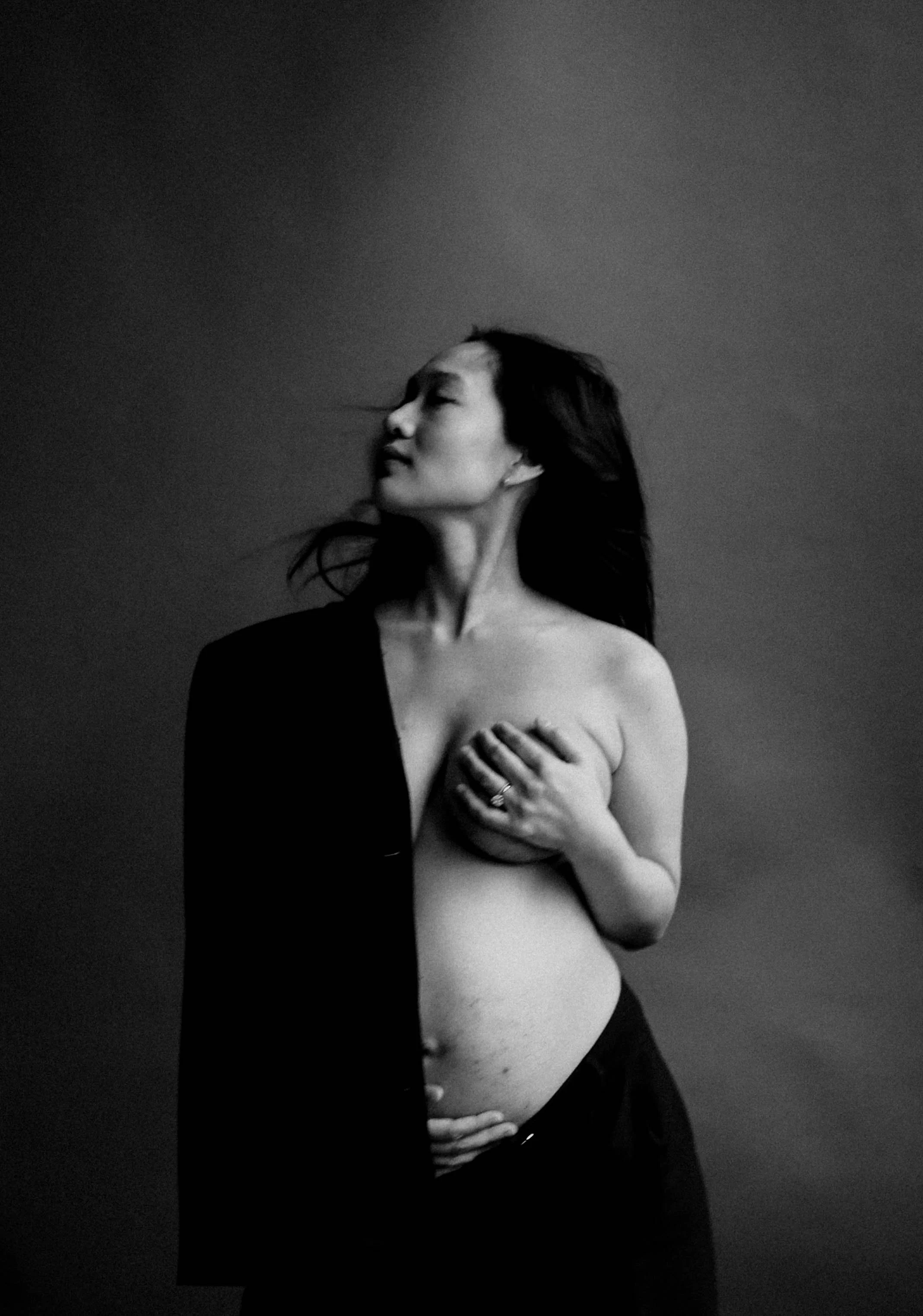 Black and white photo of a pregnant woman with Asian features, topless, covering her breast with one hand, her other hand resting on her belly, her eyes closed, and her hair flowing.