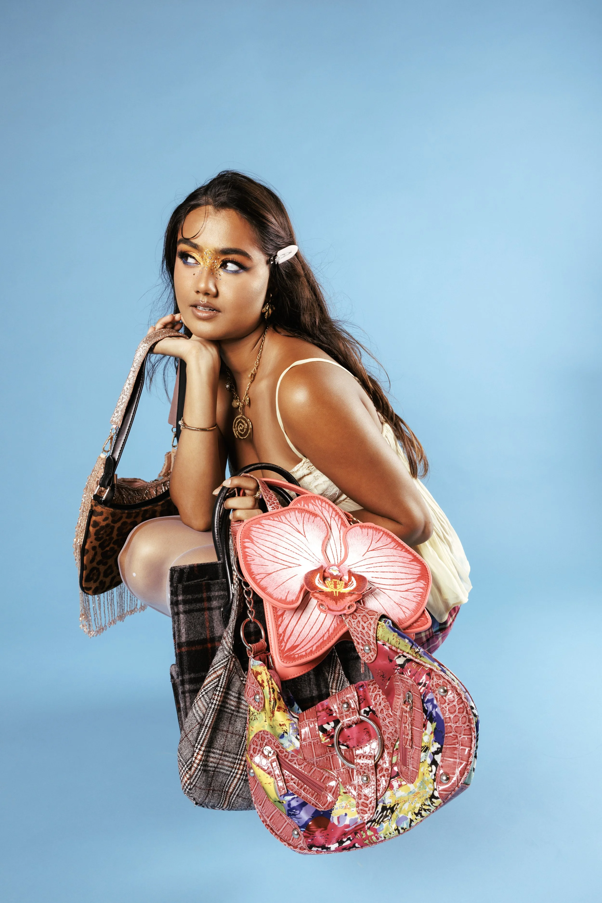 Young woman with long dark hair, wearing a cream-colored dress and colorful accessories, posing against a light blue background while holding multiple handbags and bags, including a pink floral bag and a leopard print bag.