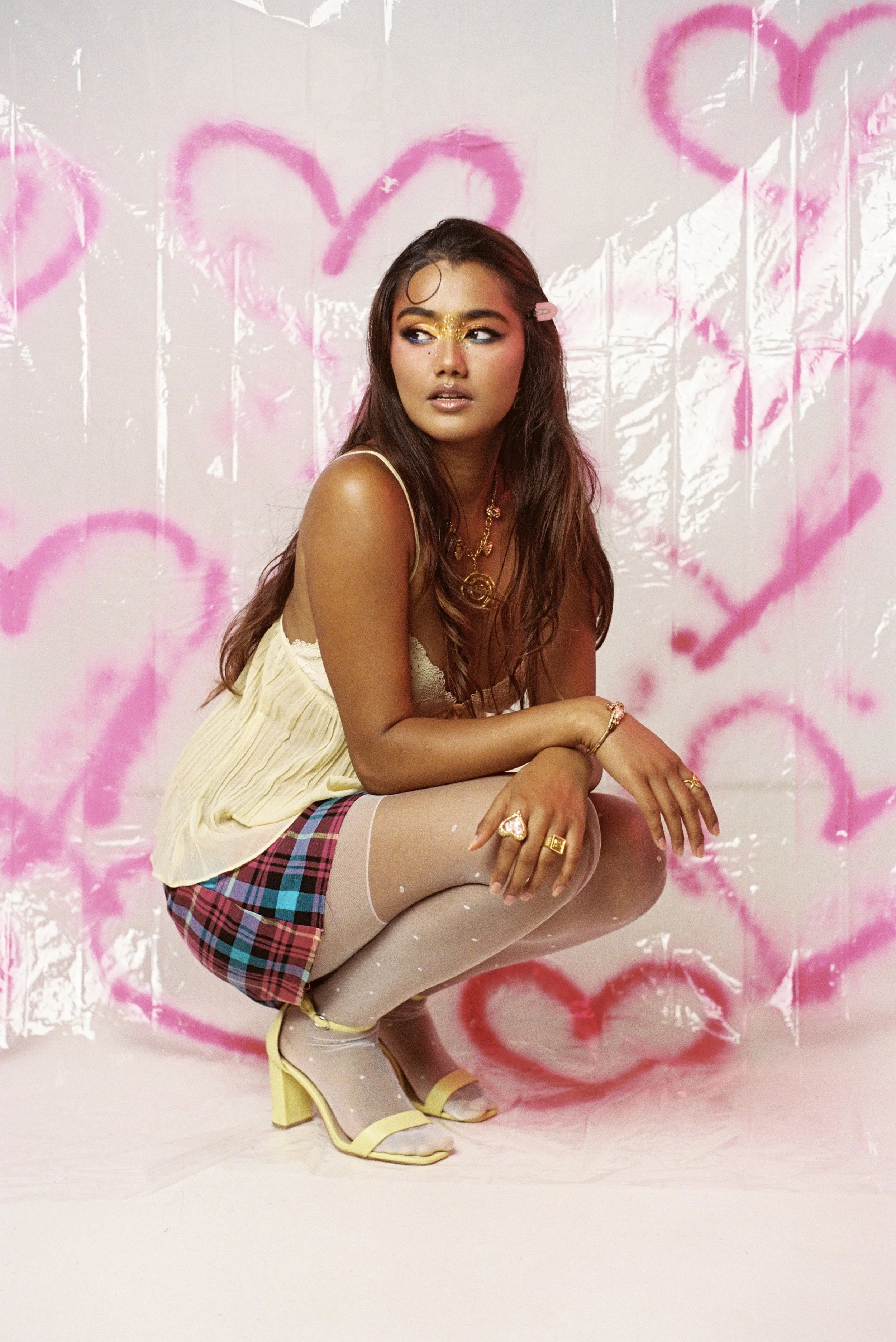 A woman squatting in front of a backdrop with pink heart graffiti, wearing a yellow top, plaid skirt, beige tights with white polka dots, and yellow high heels. She has long wavy hair, gold jewelry, and colorful makeup.