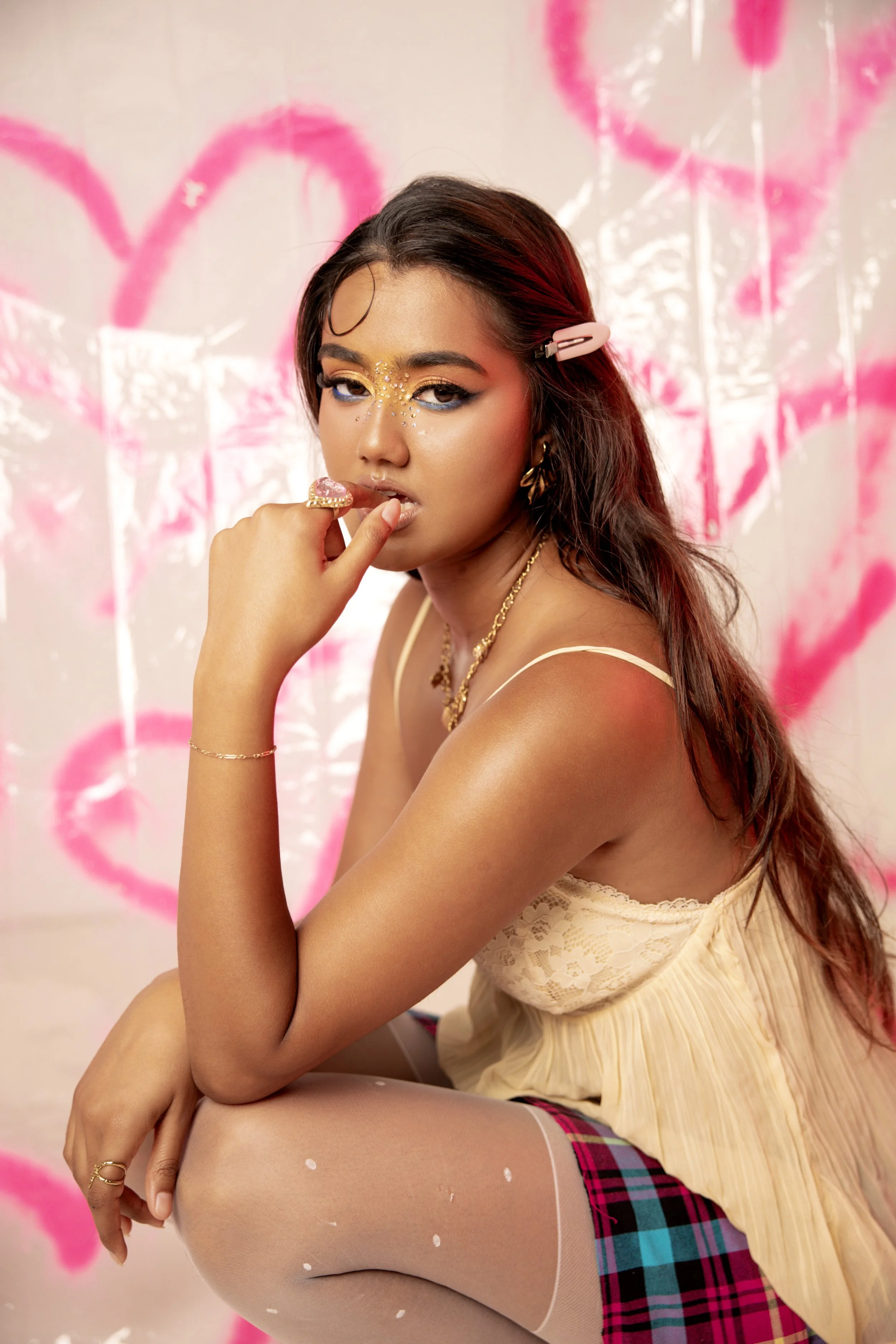 A young woman with long brown hair, wearing makeup with rhinestones around her eyes, poses with her finger near her lips. She is dressed in a beige lace top, plaid skirt, white stockings with pink and blue accents, and adorned with gold jewelry. The 