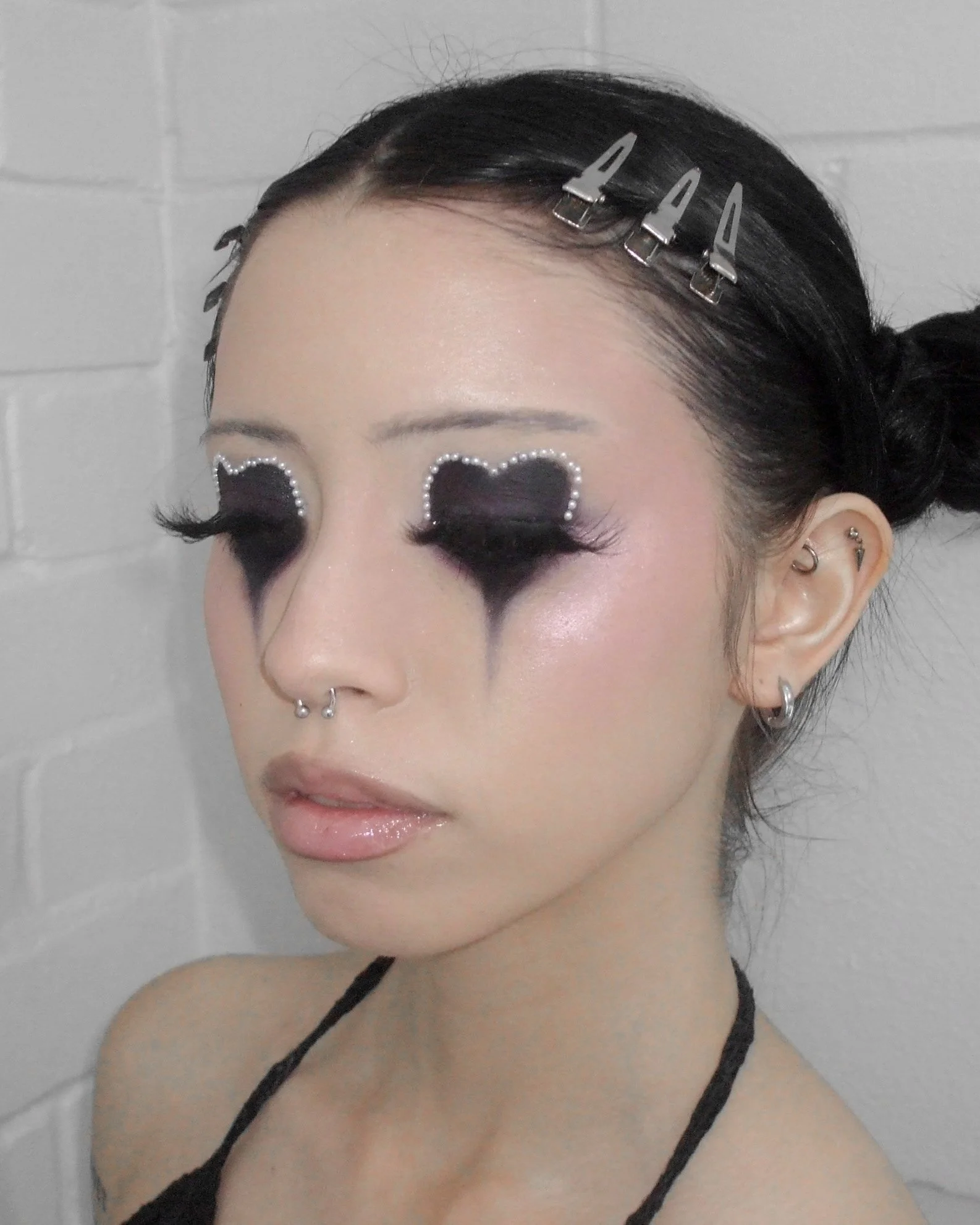Person with black and white makeup and eye decorations, wearing hair clips and earrings, against a white brick wall background.