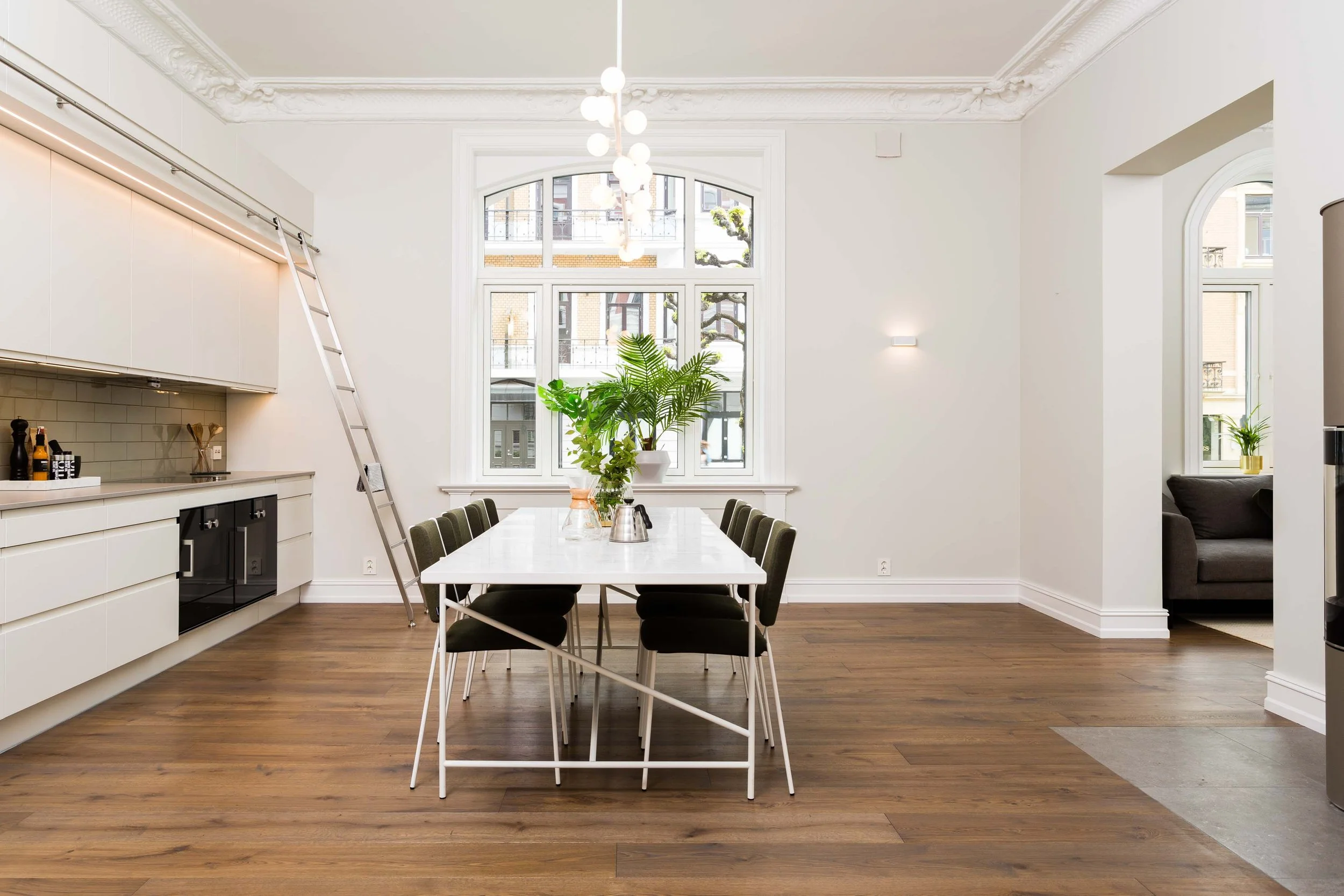 Et lyst og moderne kjøkken og spisestue med hvite vegger, stor vindu, trebord med grønne stoler, og grønne planter på bordet.