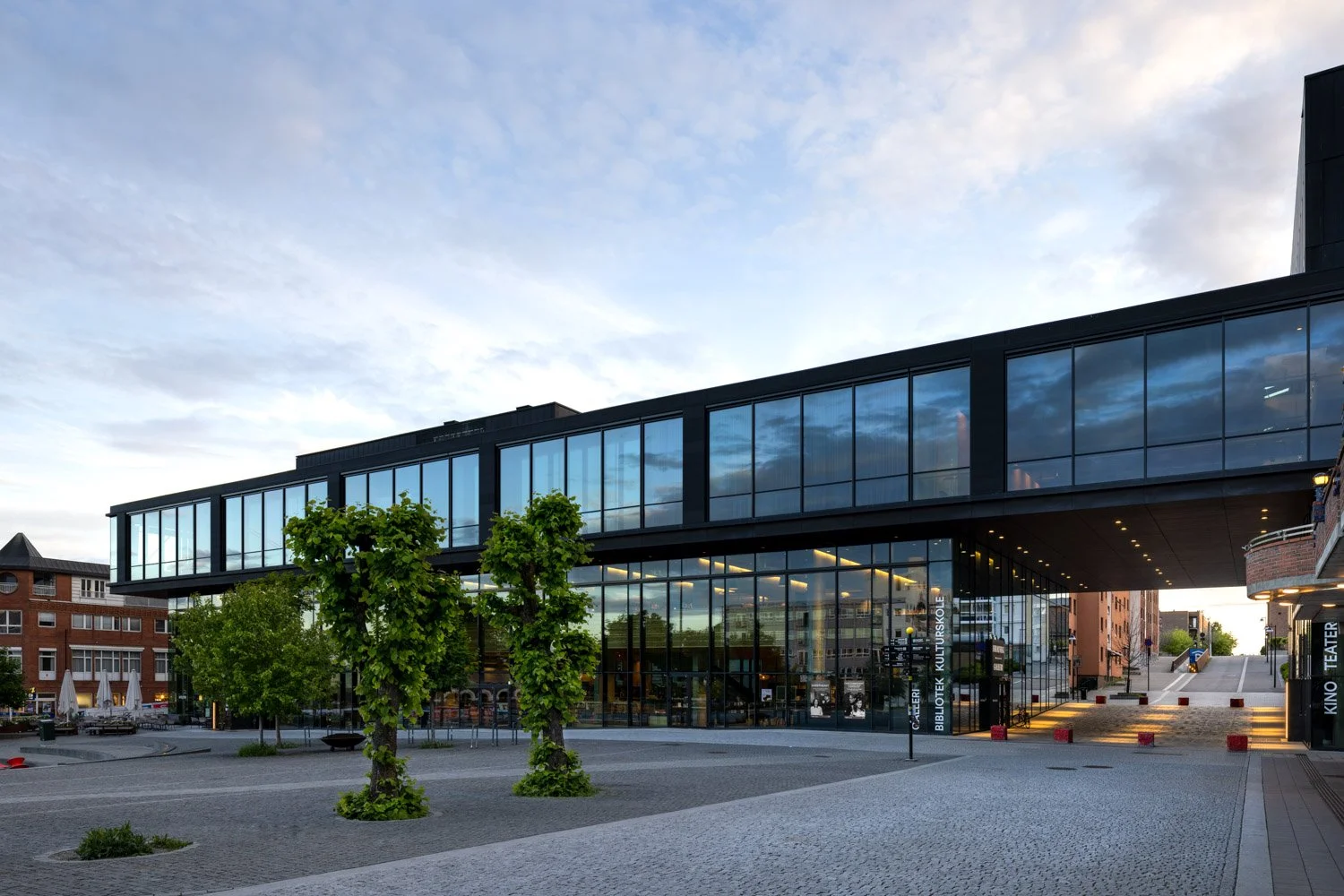 Moderne bygning med glassfasade, omgitt av trær i et byområde på kvelden.