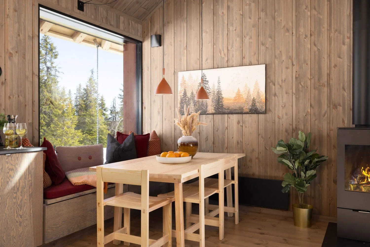 Innendørs spiseområde med trebord, fire trebarnestoler, en benk med puter, et bilde av skog på veggen, en grønn plante i gullfarget potte, og store vinduer som viser utendørs trær