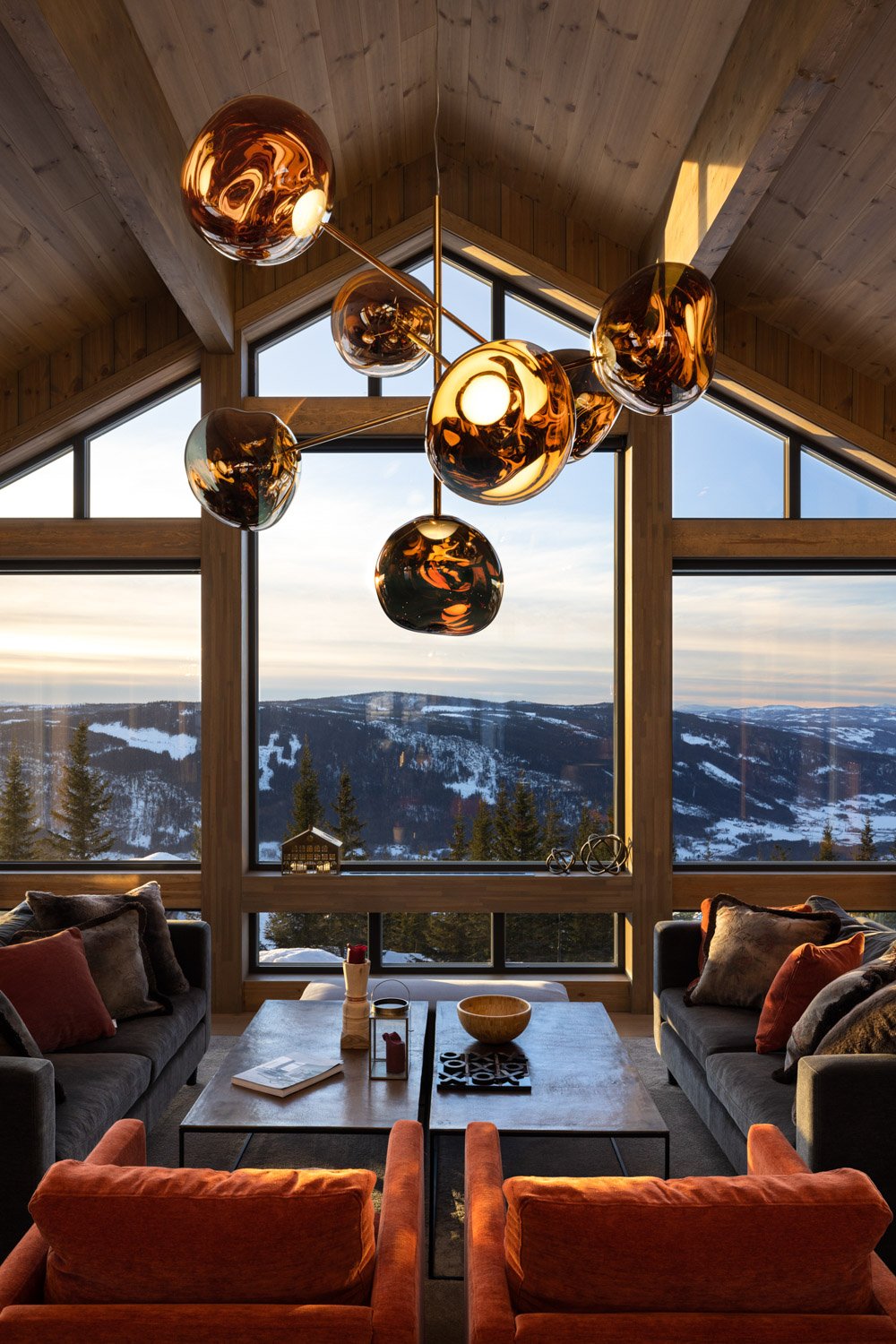Innendørs stue med store vinduer som viser snødekte fjell og skog, moderne messing lysekrone, sofagruppe med mørke og oransje puter, rustfargede lenestoler, og et bord med dekorative gjenstander.