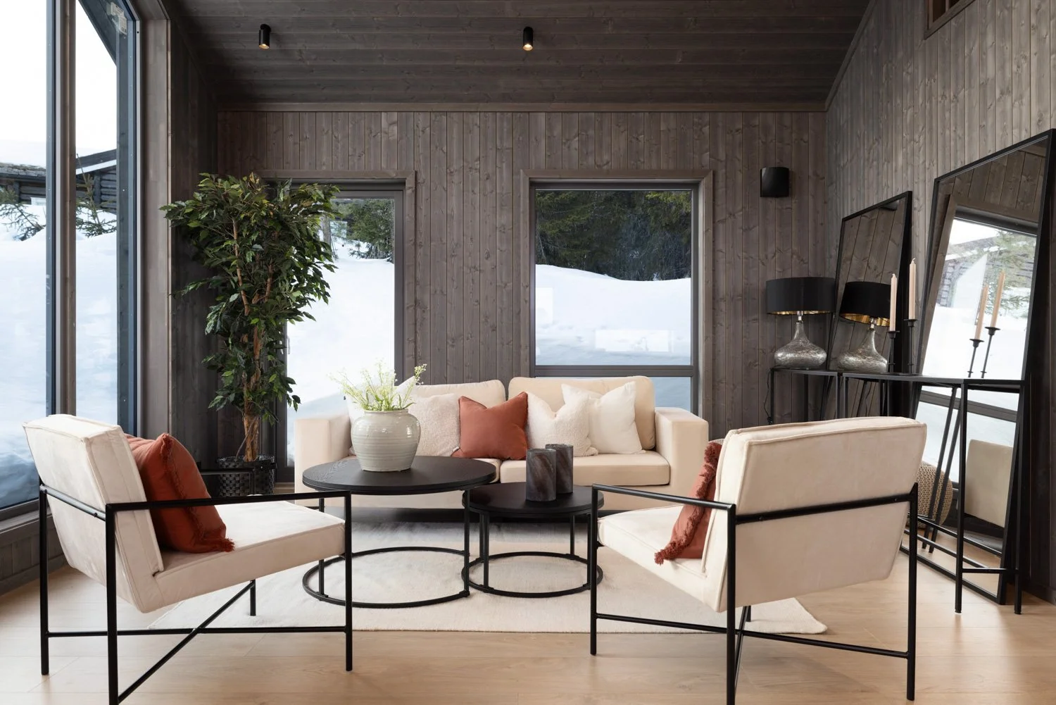 En stue med moderne møbler, inkludert en lysfarget sofa, to hvite lenestoler med rustfargede puter, et svart rundt bord, en stor plante, to store speil, og to lampebord med svarte lamper, med snø utenfor vinduene.