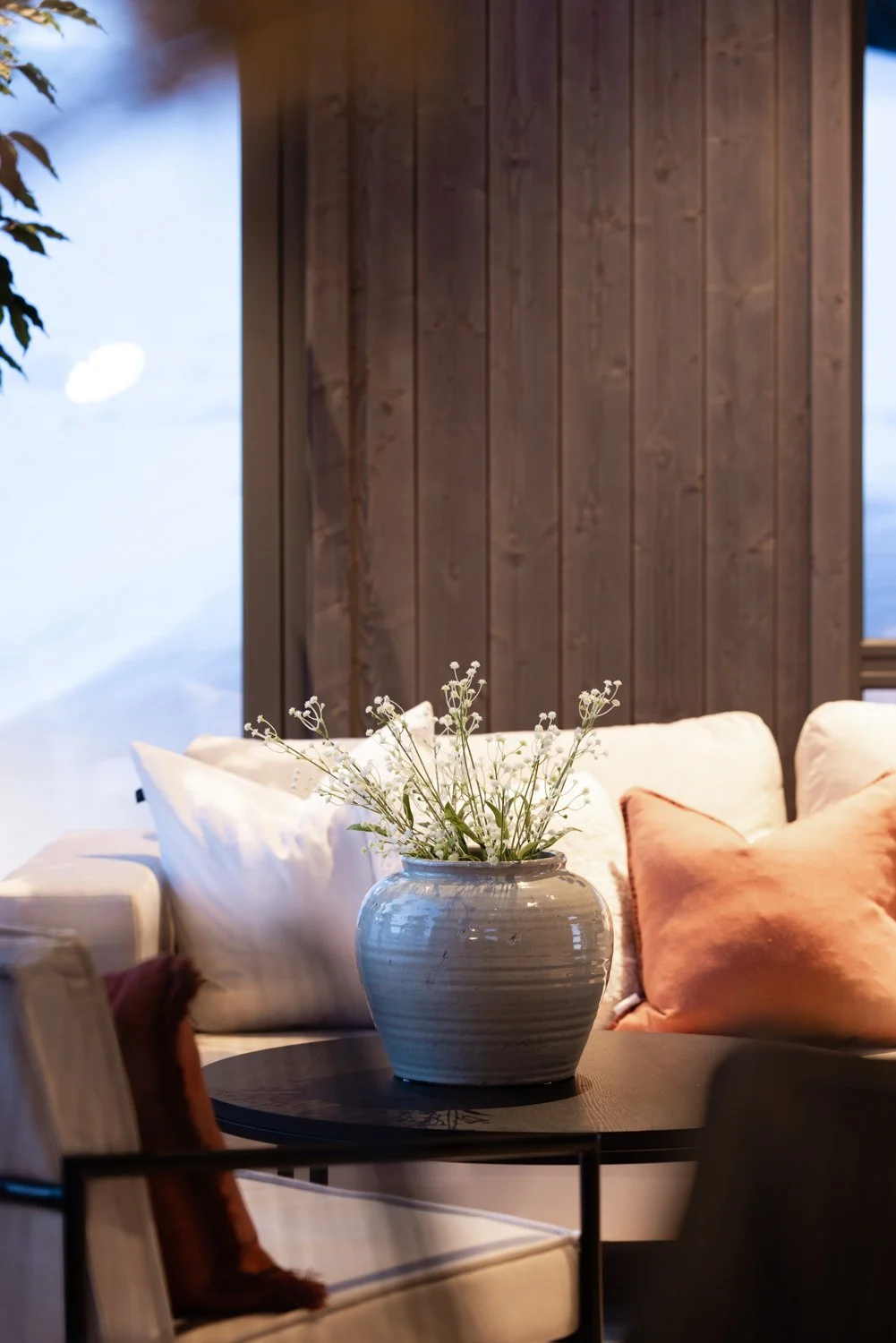 Innendørs stue med beige sofa, en hvit vase med hvite blomster på et bord, og tre puter i beige og brunt.