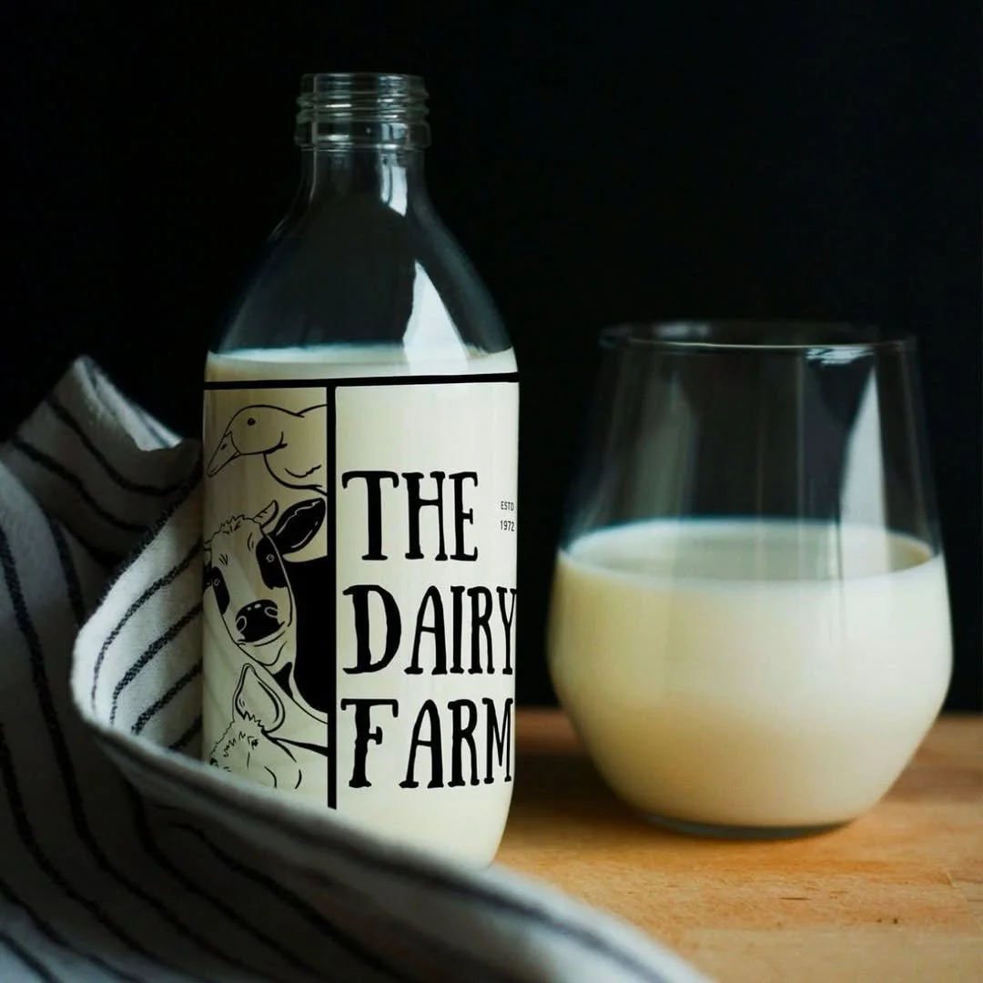 A glass bottle labeled 'The Dairy Farm' with illustrations of farm animals, partially filled with milk, next to a glass of milk on a wooden surface, with a striped cloth.