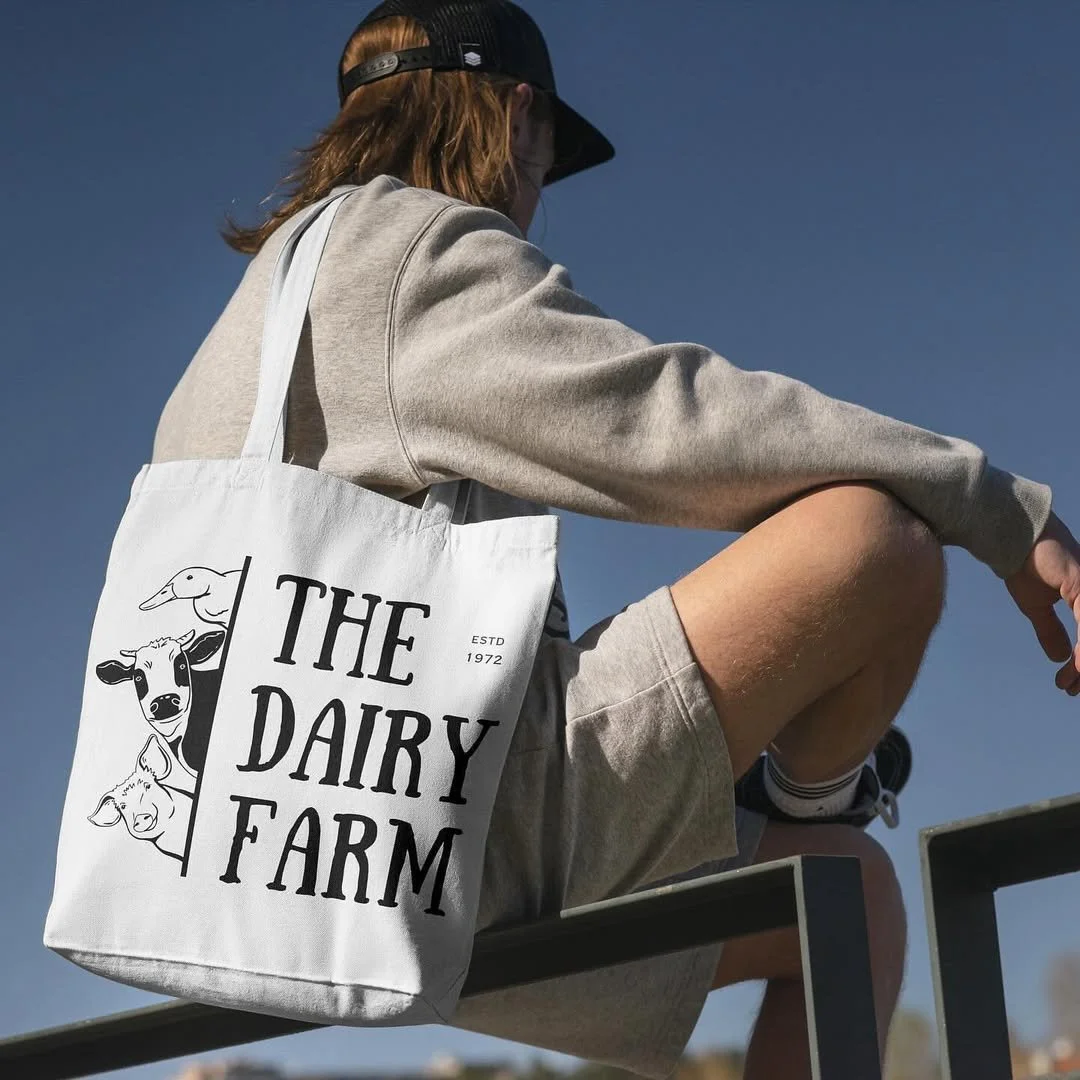 A person wearing a beige sweatshirt, shorts, and a black hat sitting on a railing outdoors, carrying a white tote bag with 'THE DAIRY FARM' text and illustrations of farm animals against a clear blue sky.
