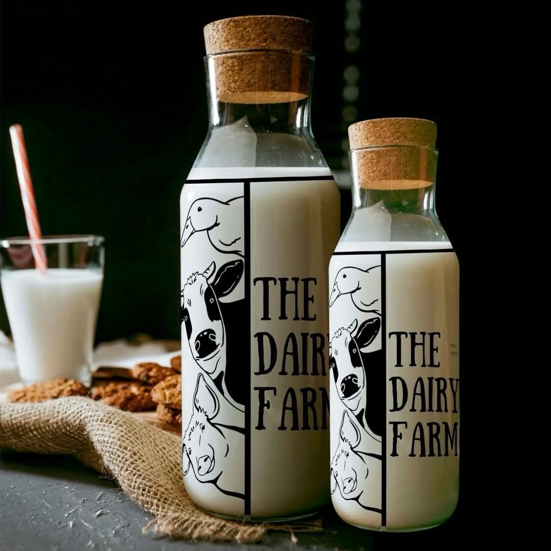 Two glass bottles labeled "The Dairy Farm" with cork stoppers, filled with milk, set on a table with a glass of milk, cookies, and a burlap cloth, with a dark background.