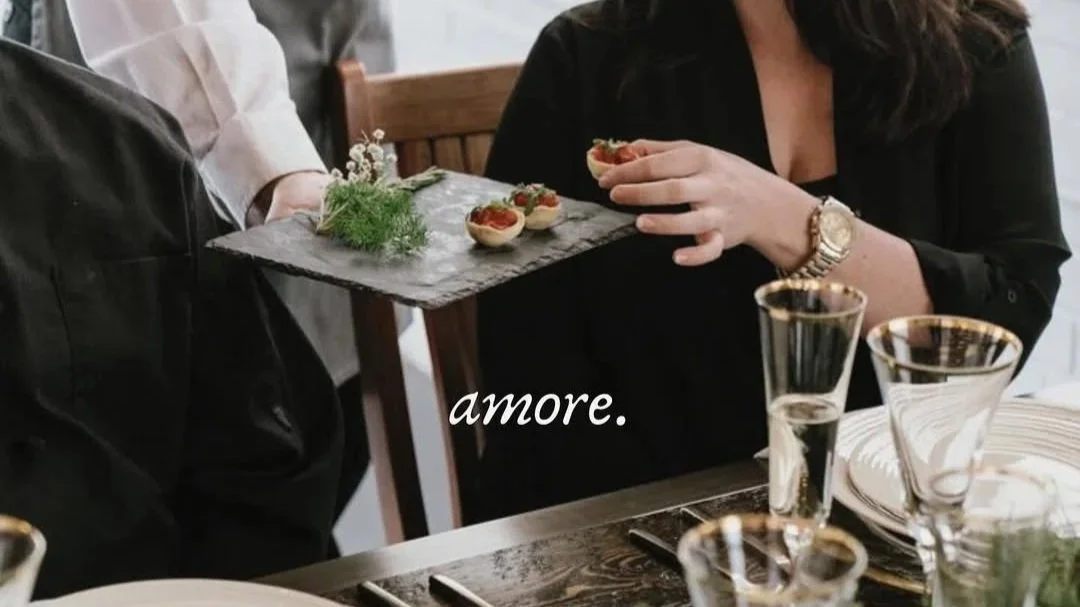 A woman dressed in black holding a hand of small pasta dishes on a stone serving tray, with a table set for a meal including wine glasses and plates, and the word 'amore' written on the image.