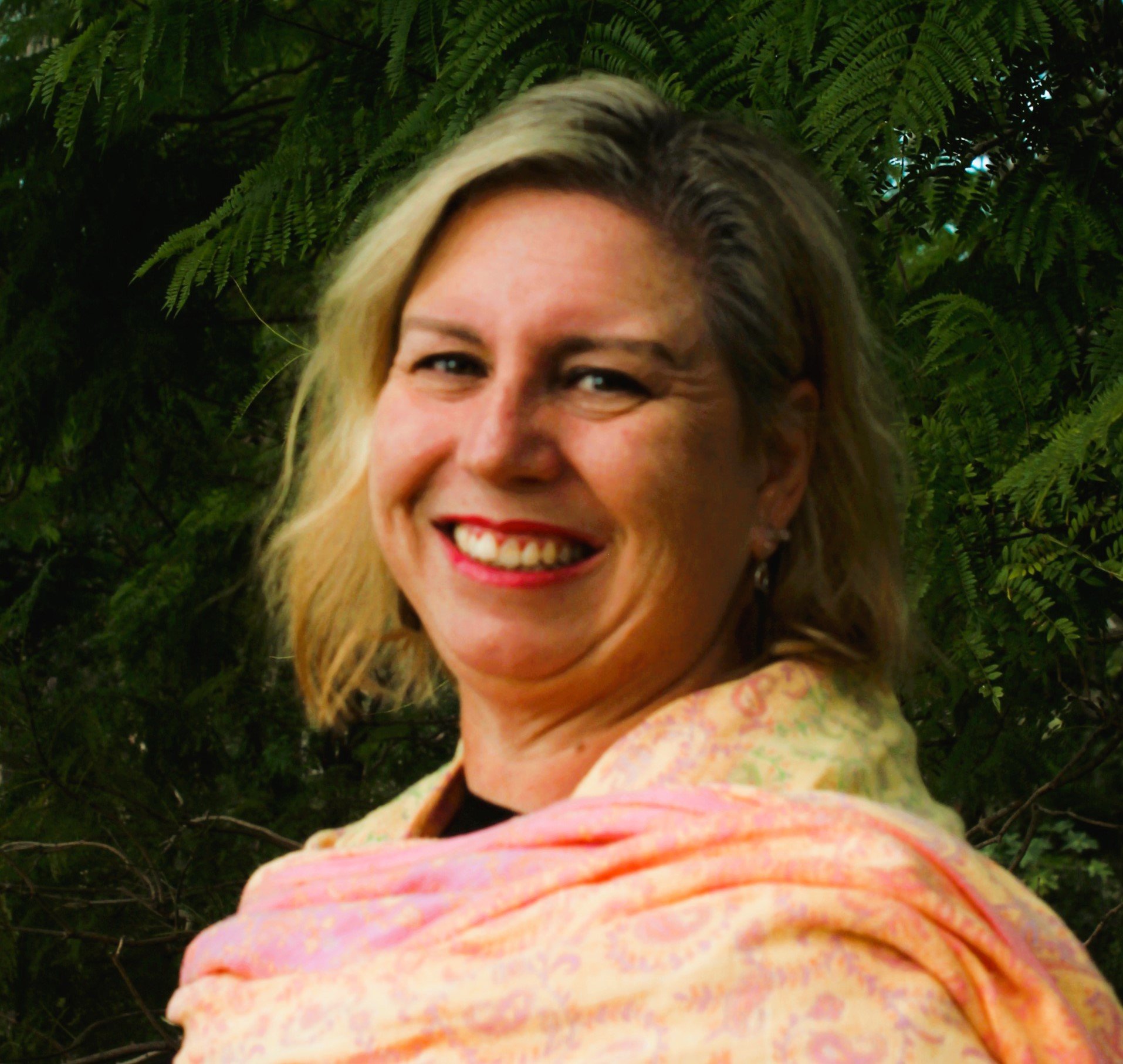 Close-up of a smiling woman with shoulder-length blond hair, wearing a pastel-colored shirt, standing in front of green fern leaves.