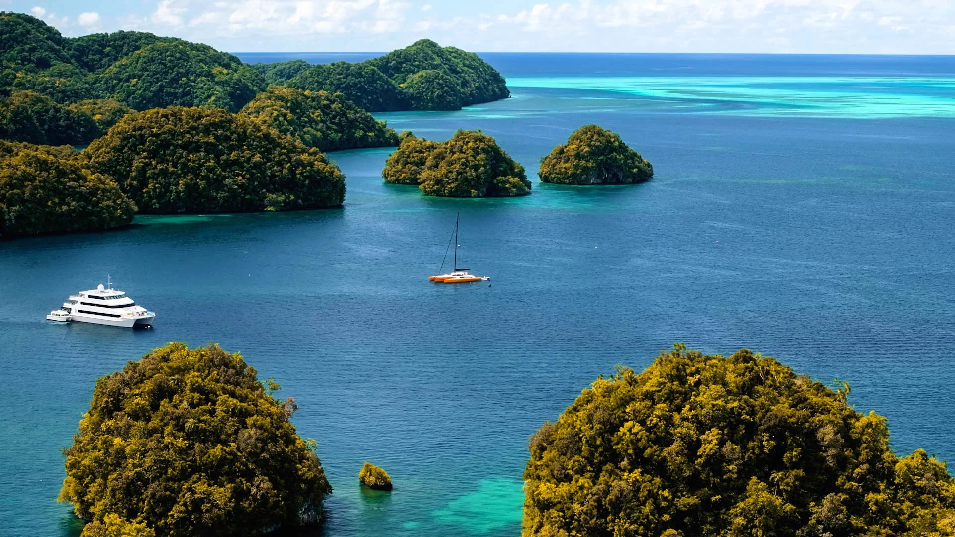 A scenic view of clear blue ocean water with lush green islands and three boats, including a large yacht and two smaller boats, floating near the islands.