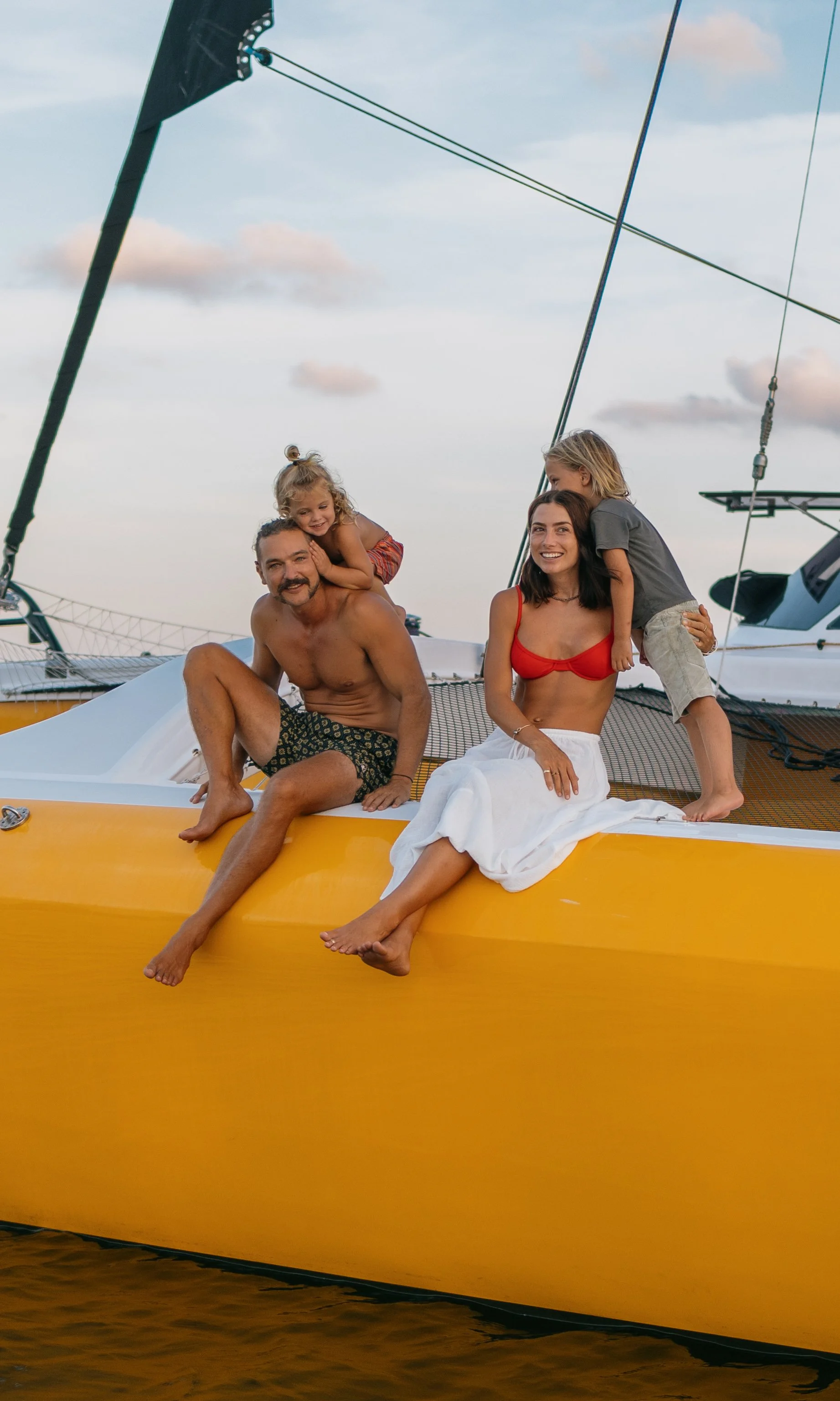 A family of four sitting on a yellow sailboat, smiling, with a blue sky and clouds in the background.