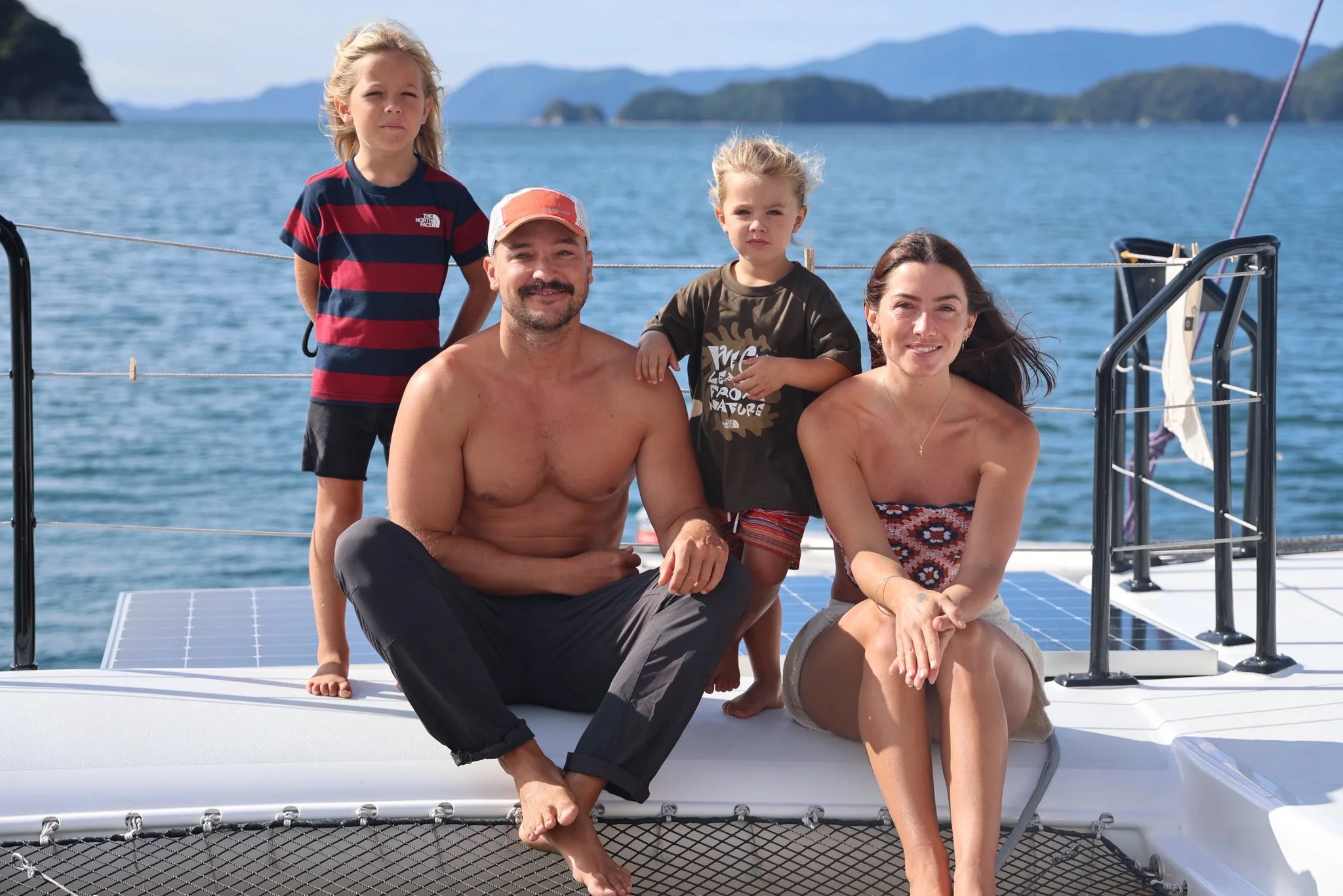 A family of four sitting on the dock of a boat with a scenic lake view and mountains in the background. The father is shirtless, wearing a cap, and sitting cross-legged. The mother is sitting with her legs crossed, wearing a strapless top. The two children, a girl and a boy, are standing behind their parents, looking towards the camera. The girl is wearing a striped T-shirt, and the boy is wearing a T-shirt with a graphic design. The family appears relaxed and enjoying their time on the water.