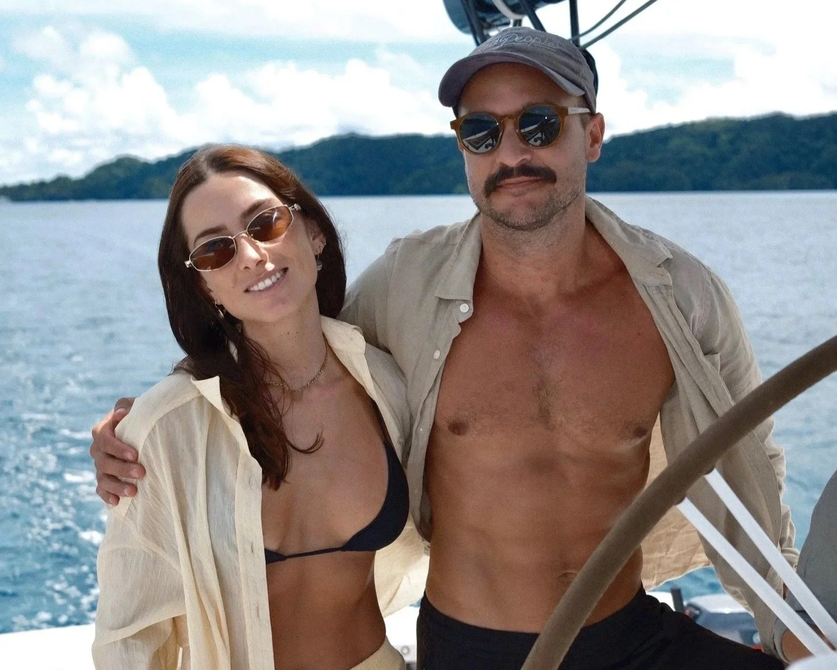 A man and woman posing on a boat with a water and island background, both wearing sunglasses and swimwear, with the man shirtless and the woman in a black bikini top and open beige shirt.
