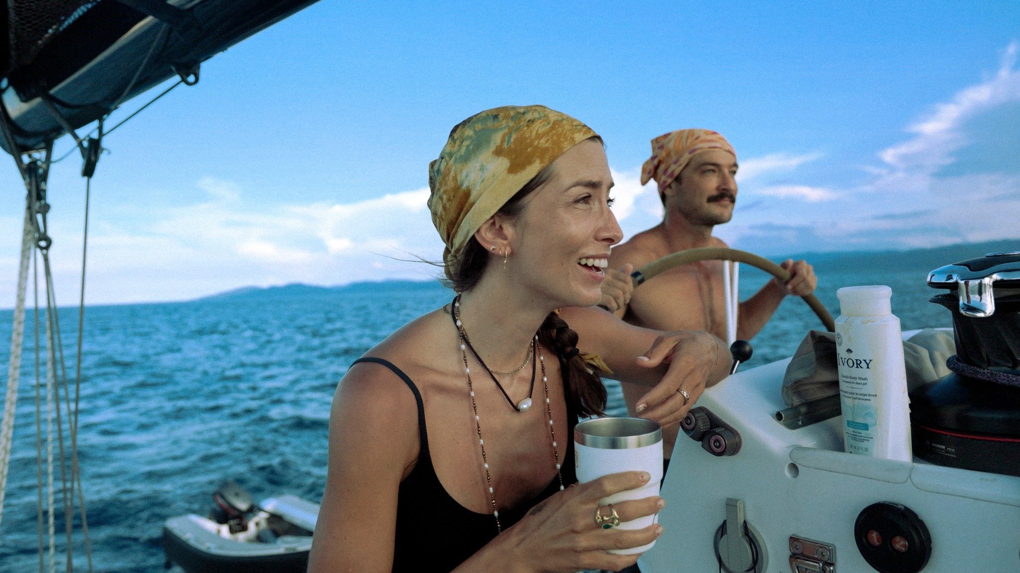 A woman with long dark hair wearing a headscarf and black dress sitting on a boat, smiling and drinking from a cup. A man with a headscarf and mustache is in the background steering the boat. The sea and sky are visible in the background.