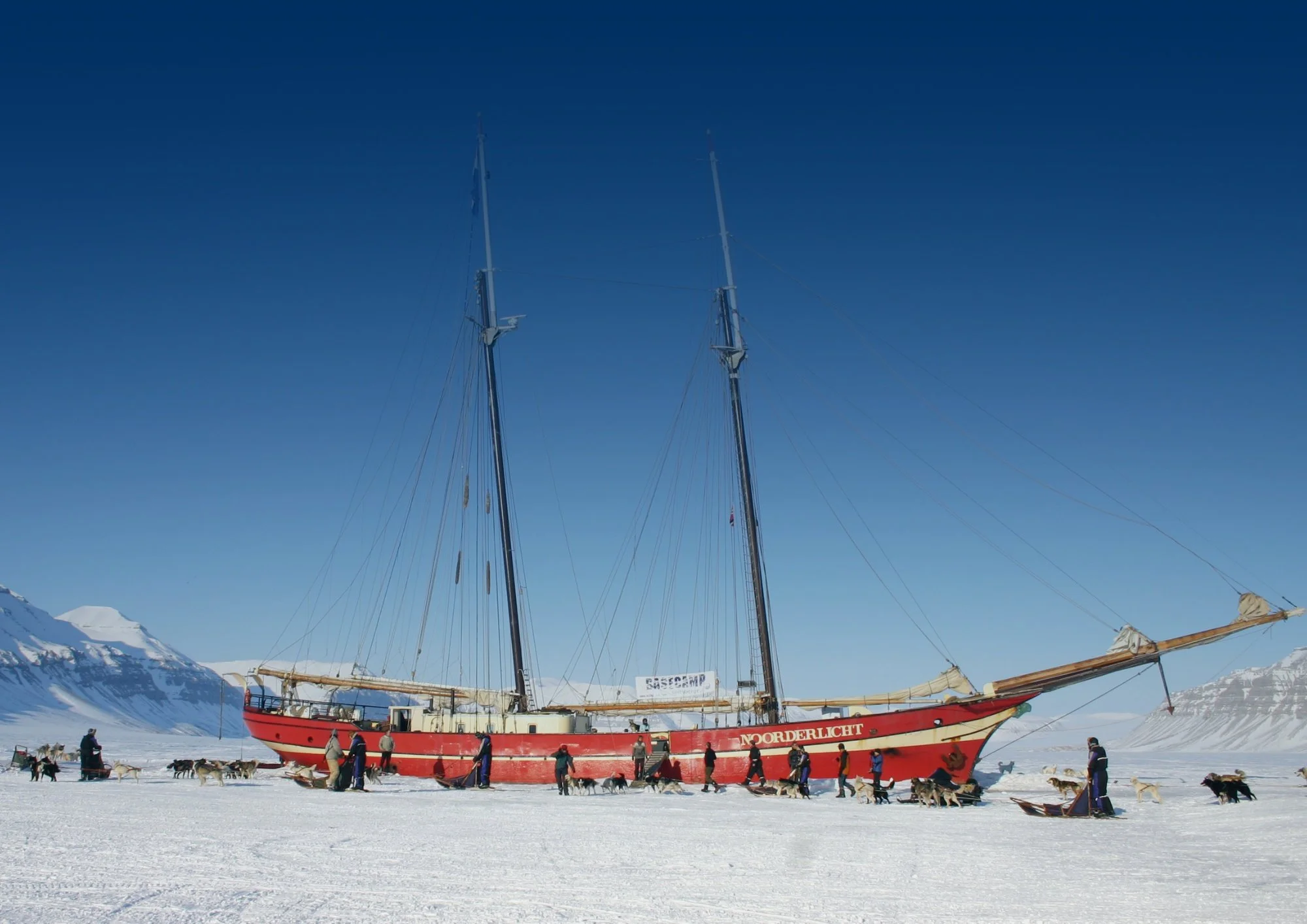 A large red and white ship named 'Norderlicht' on a snow-covered surface in a cold, mountainous area with people and sled dogs nearby.