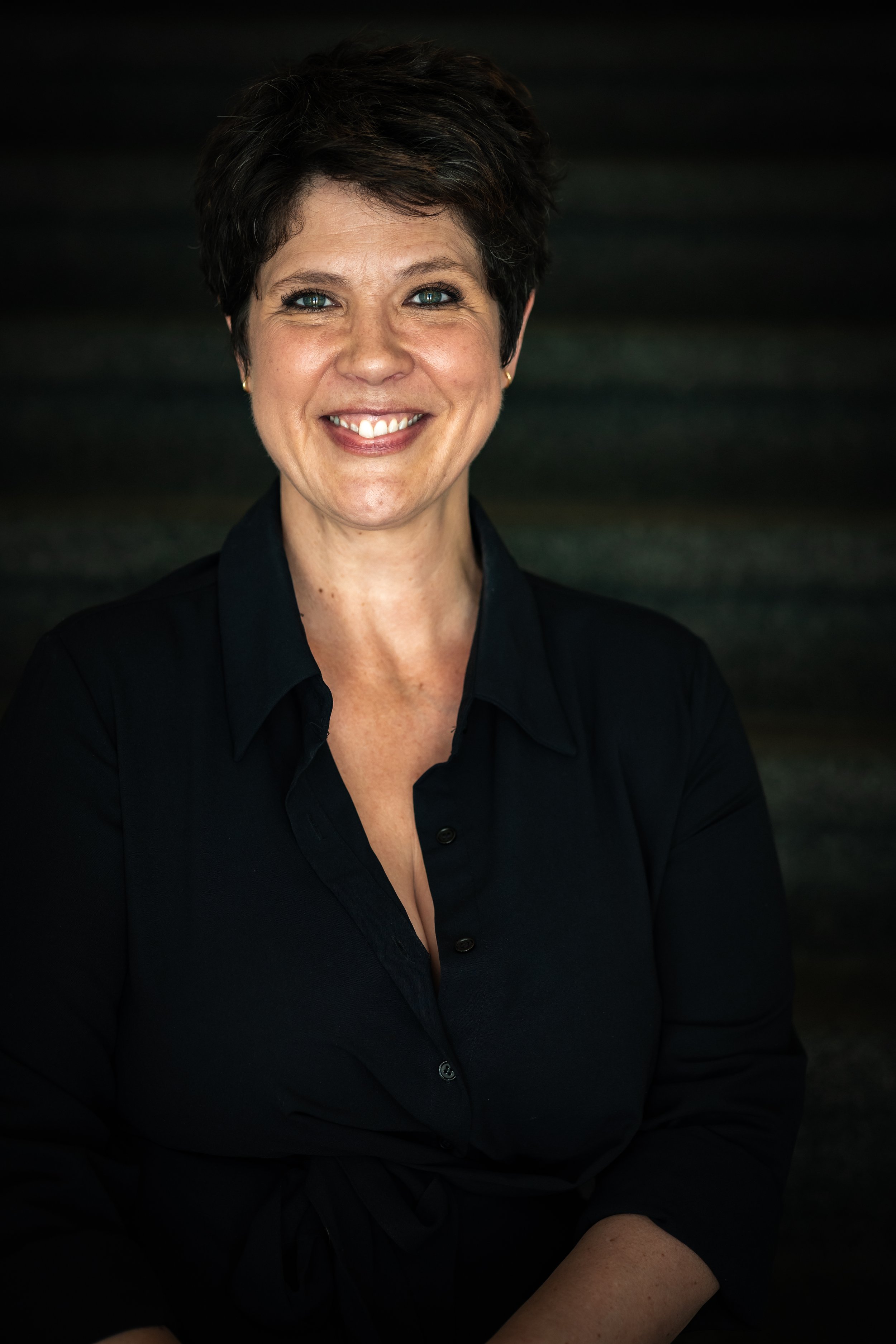 A woman with short dark hair, wearing a black collared shirt, smiling in front of a dark background.