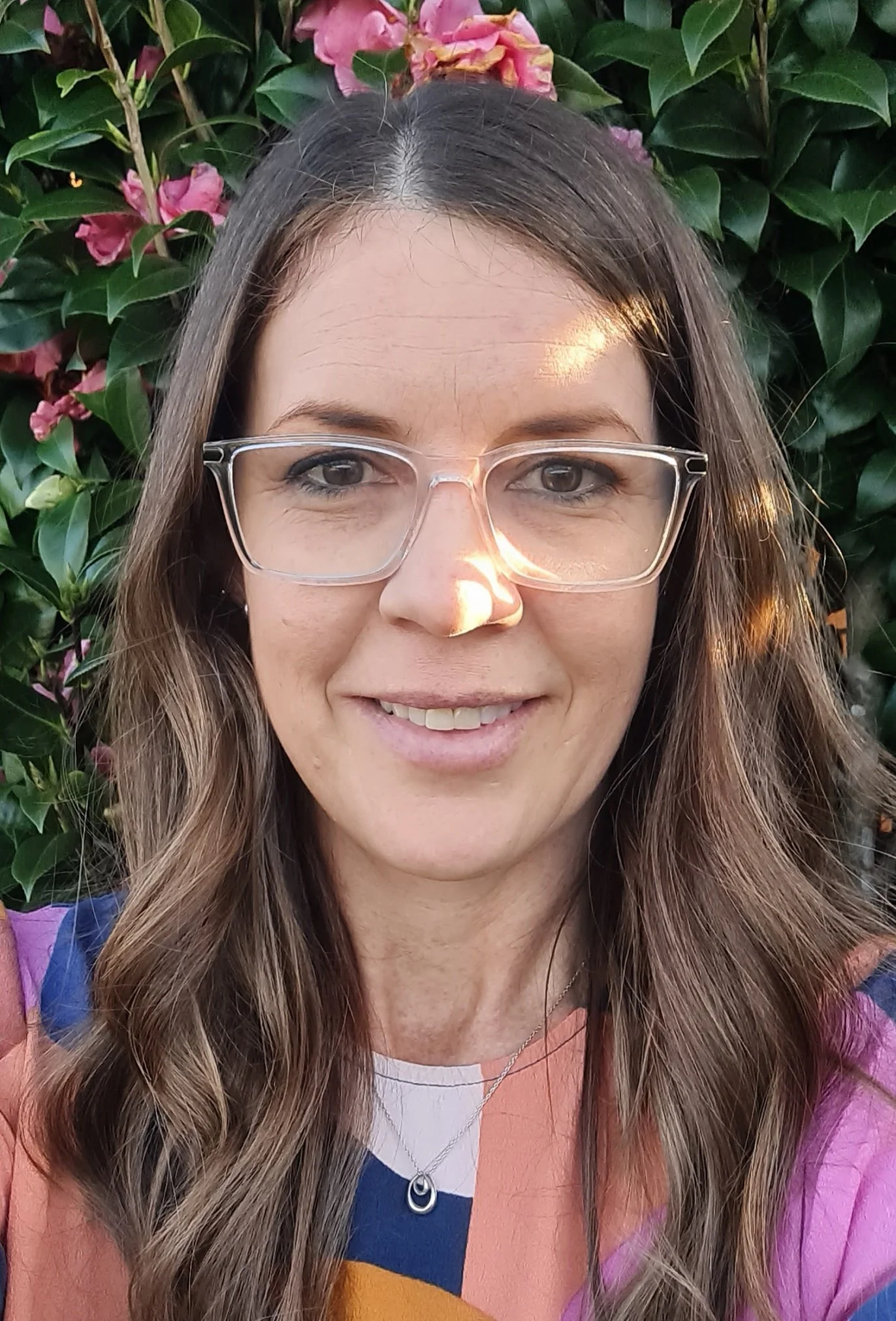 Close-up of a smiling woman with glasses, long wavy hair, and a colorful top, standing in front of green leafy plants and pink flowers.